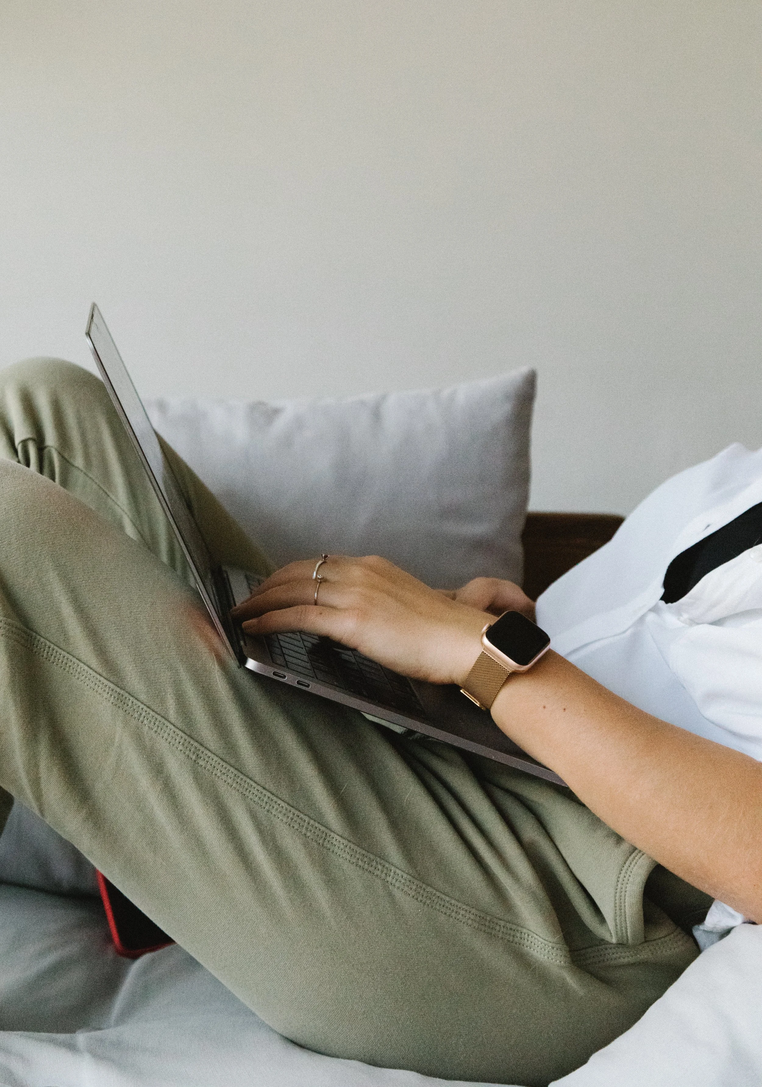 Person lying on bed using a laptop, wearing a smartwatch and a ring, with a gray pillow in the background.