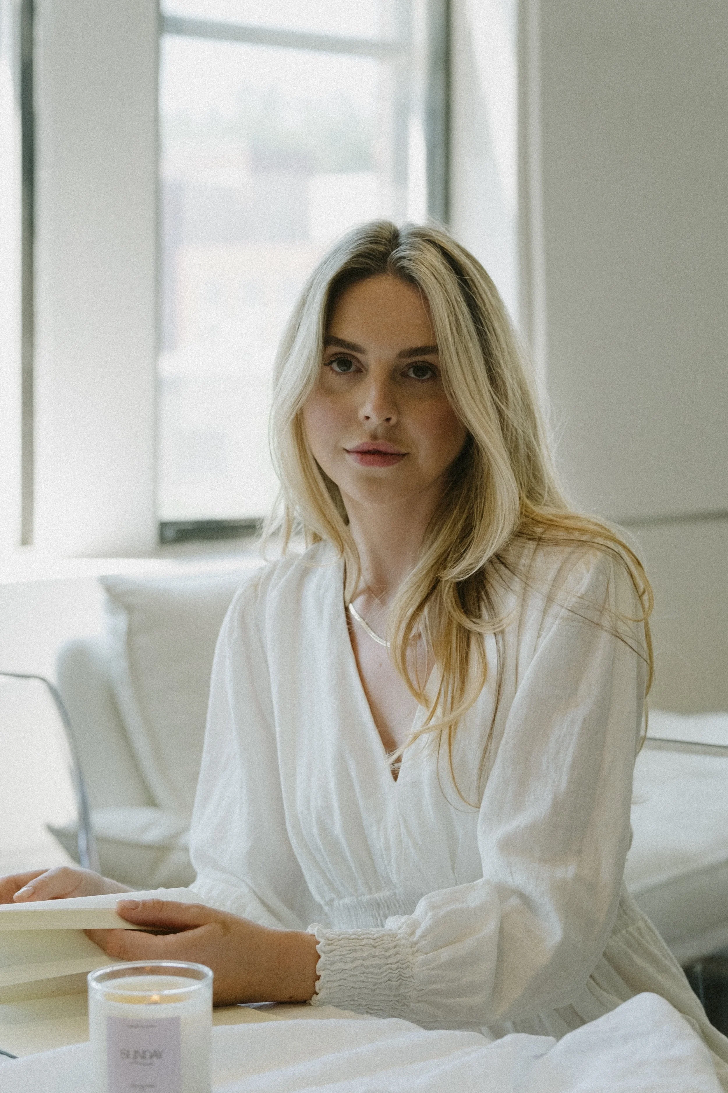 A woman with long blonde hair sitting in a well-lit room near a window, wearing a white blouse, looking at the camera.