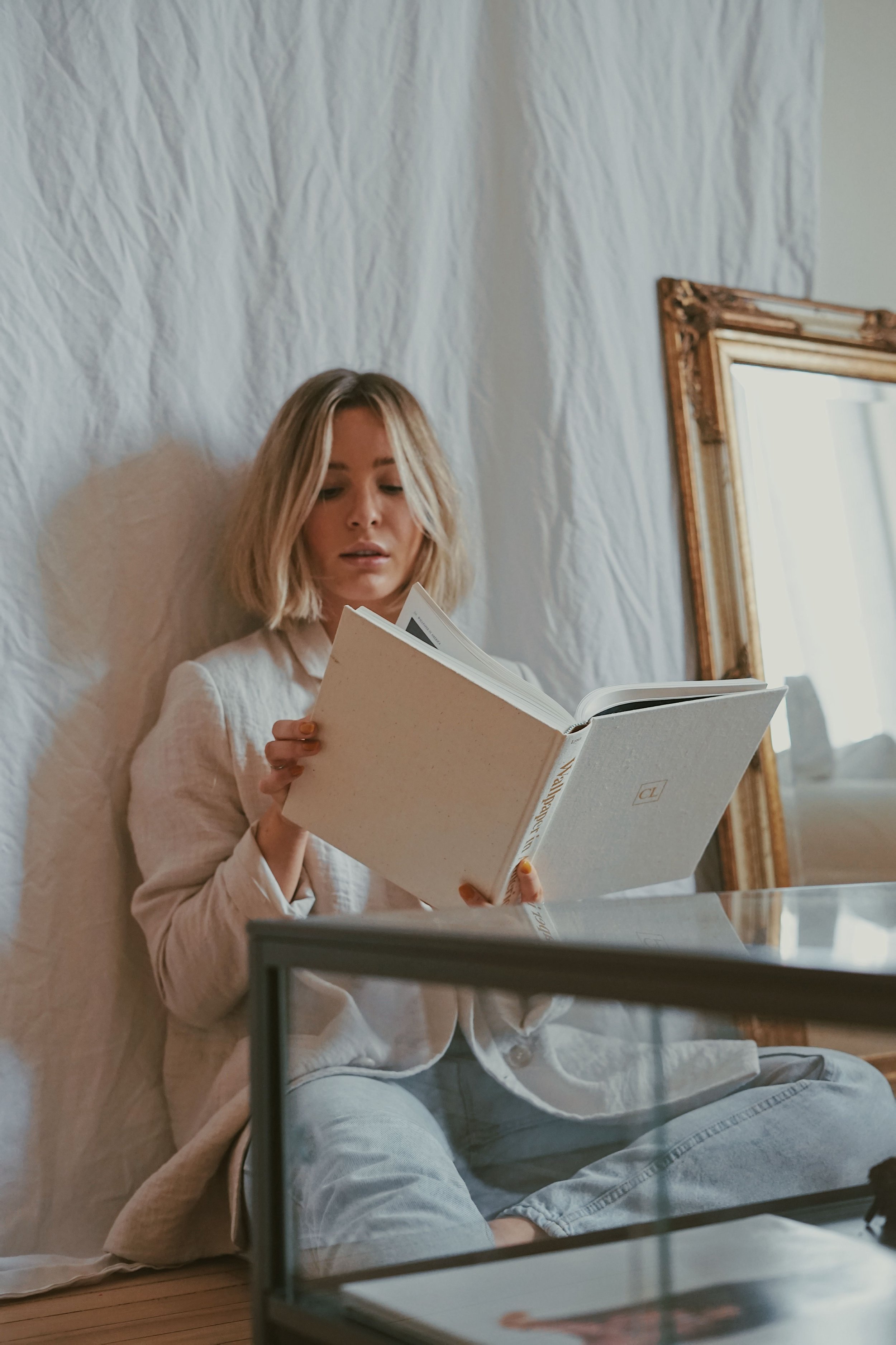 A young woman with blonde hair reading a book while sitting on the floor next to a glass display case and a mirror, with a white textured wall behind her.