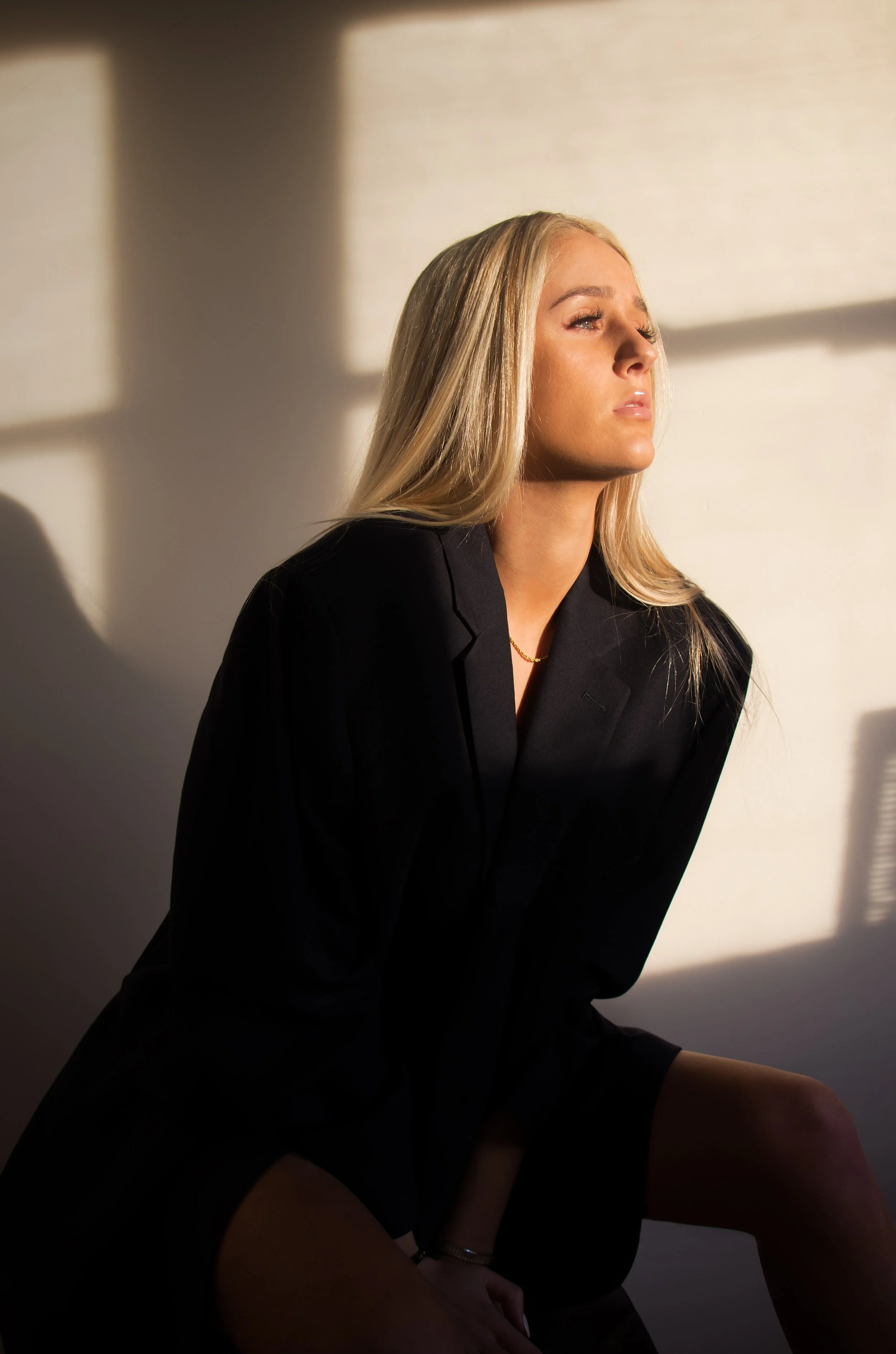 A woman with long blonde hair, dressed in a black blazer, sitting in sunlight casting shadows on the wall.