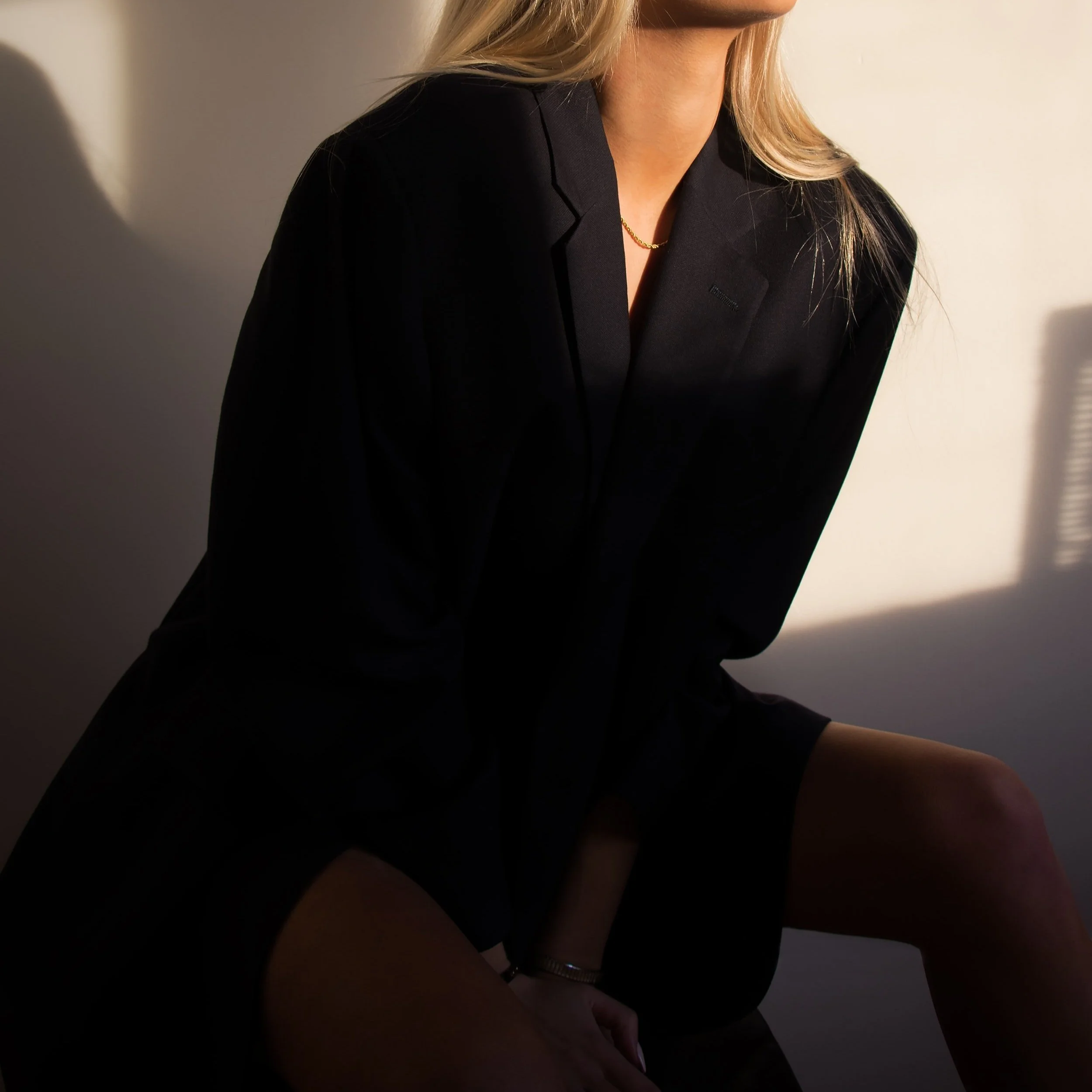 A woman with blonde hair wearing a black blazer, sitting in a room with sunlight casting shadows on the wall.