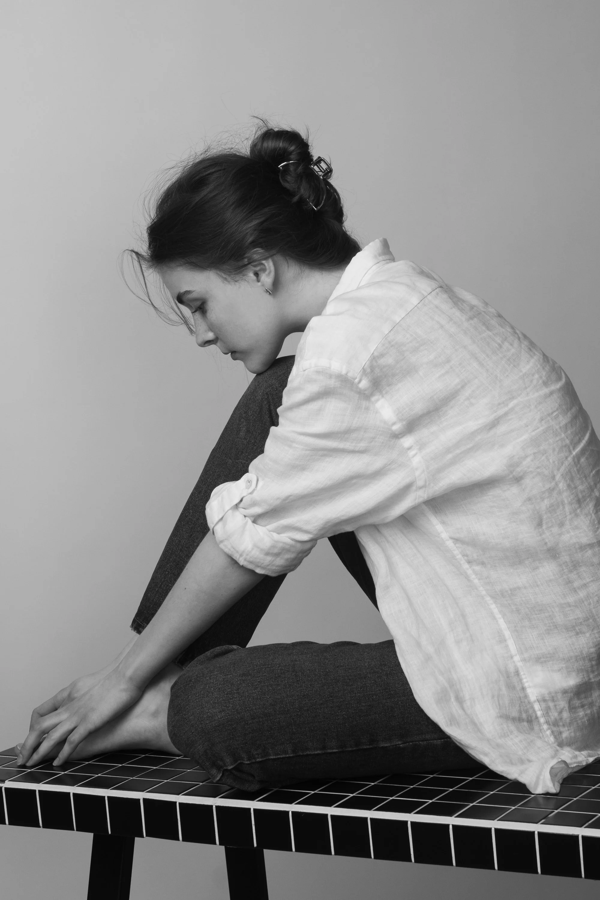 A black and white photo of a woman sitting cross-legged on a black-tiled table, stretching forward with her arms extended.