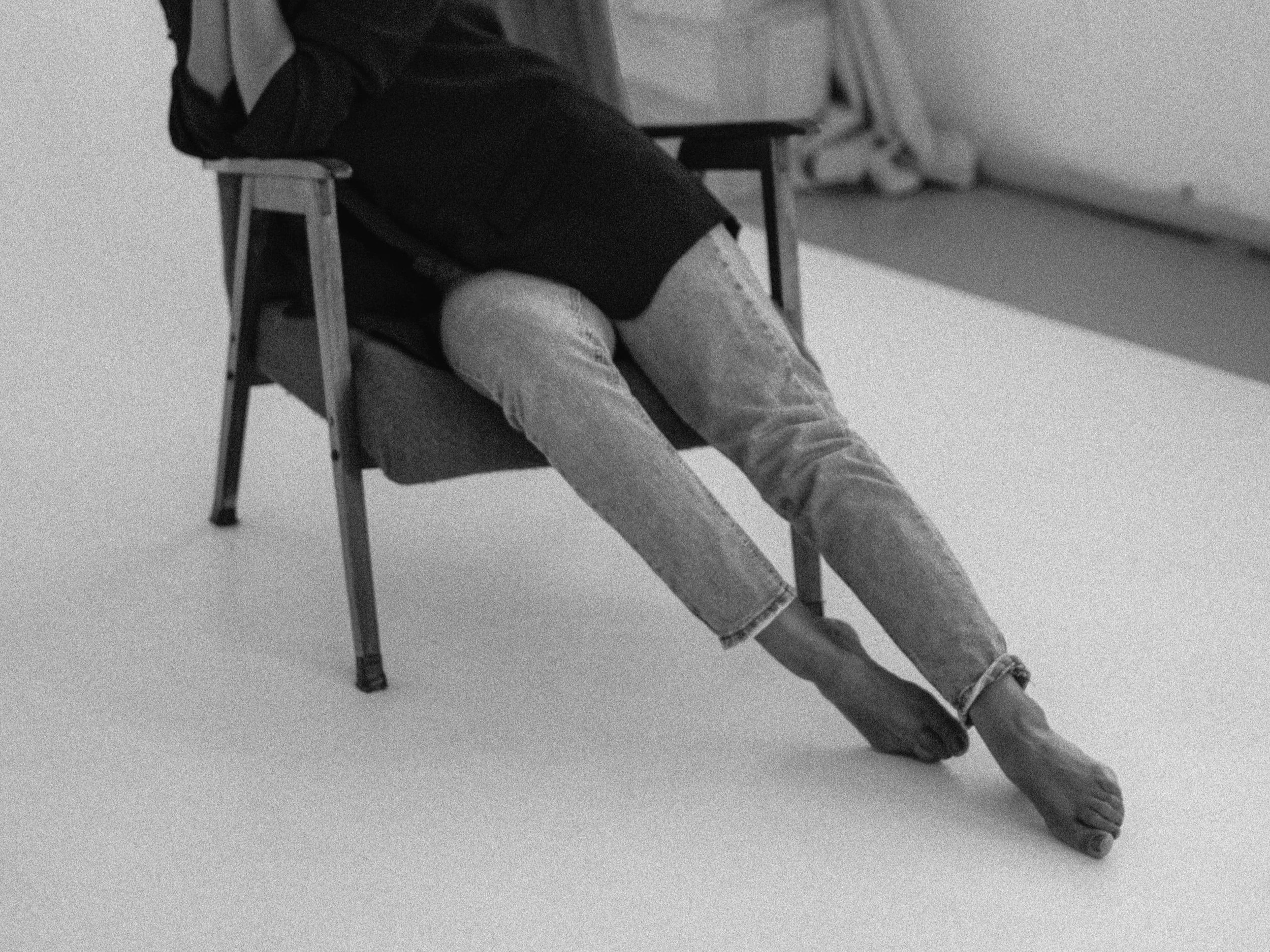 Person sitting on a wooden chair with legs crossed at the knees, dressed in a dark jacket and light jeans, with bare feet, on a plain white background.