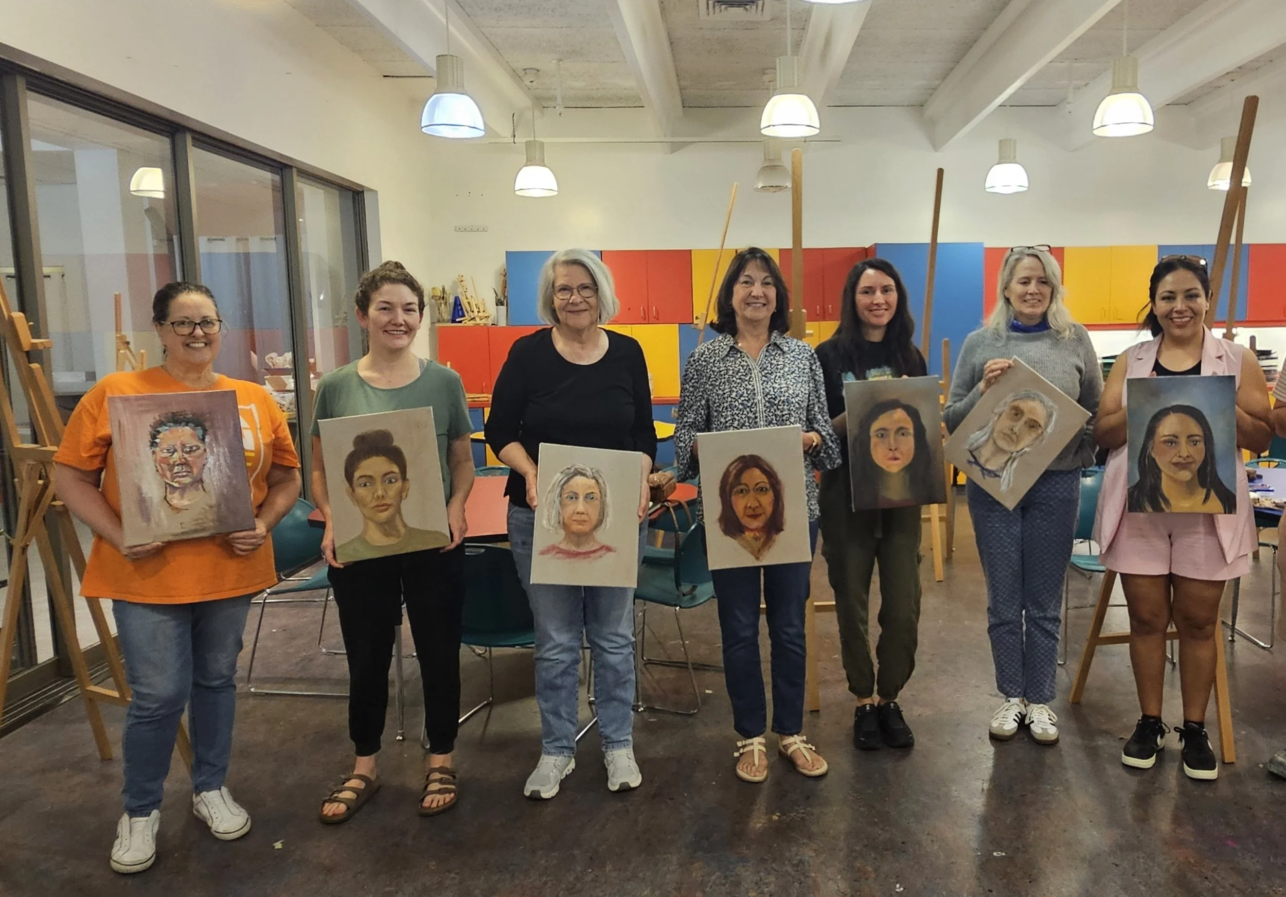 A group of seven women standing in an art classroom holding portrait paintings they created, with easels and colorful cabinets in the background.