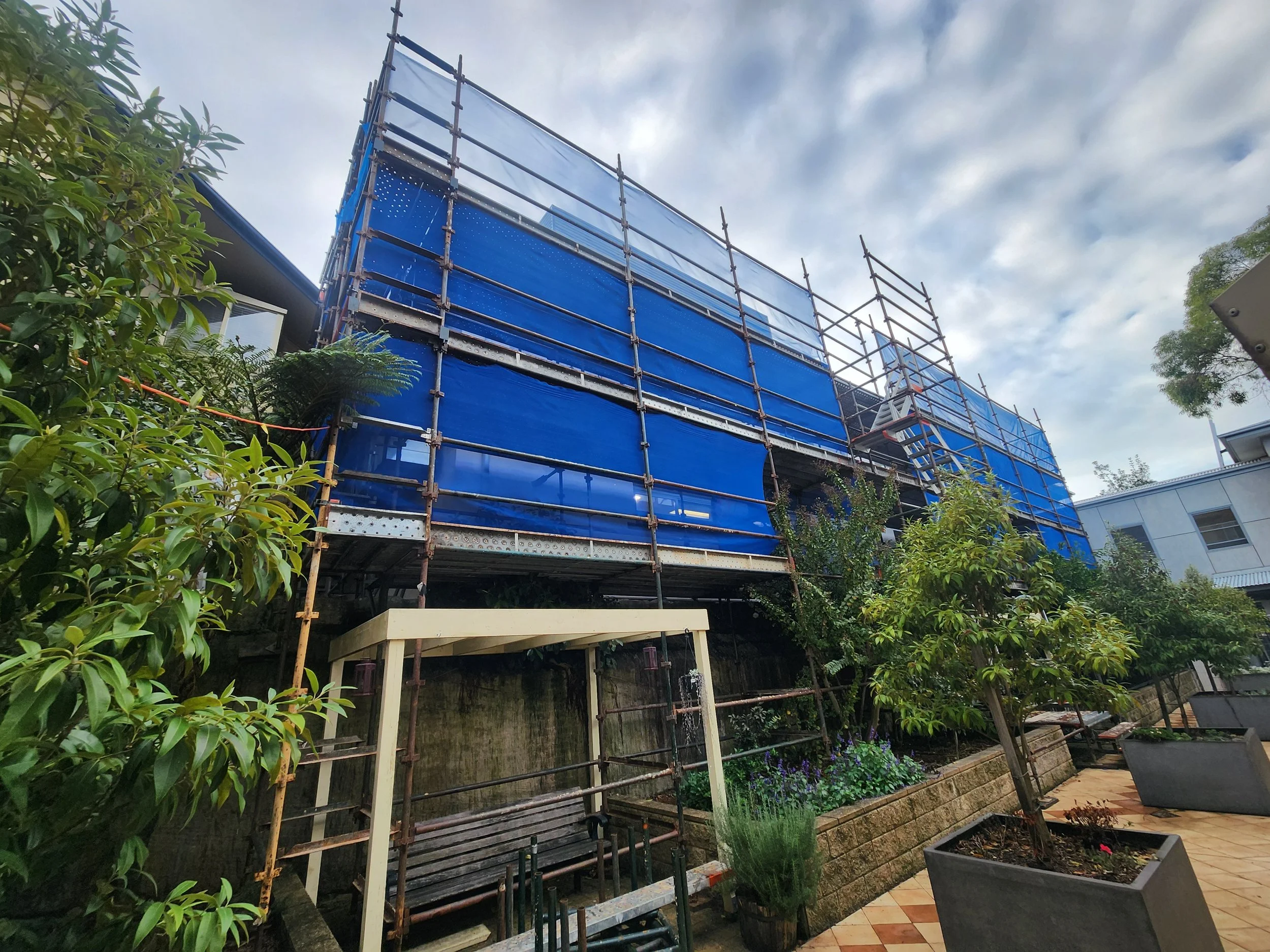 Construction scaffolding around a blue building under cloudy sky, with greenery and a garden in the foreground.