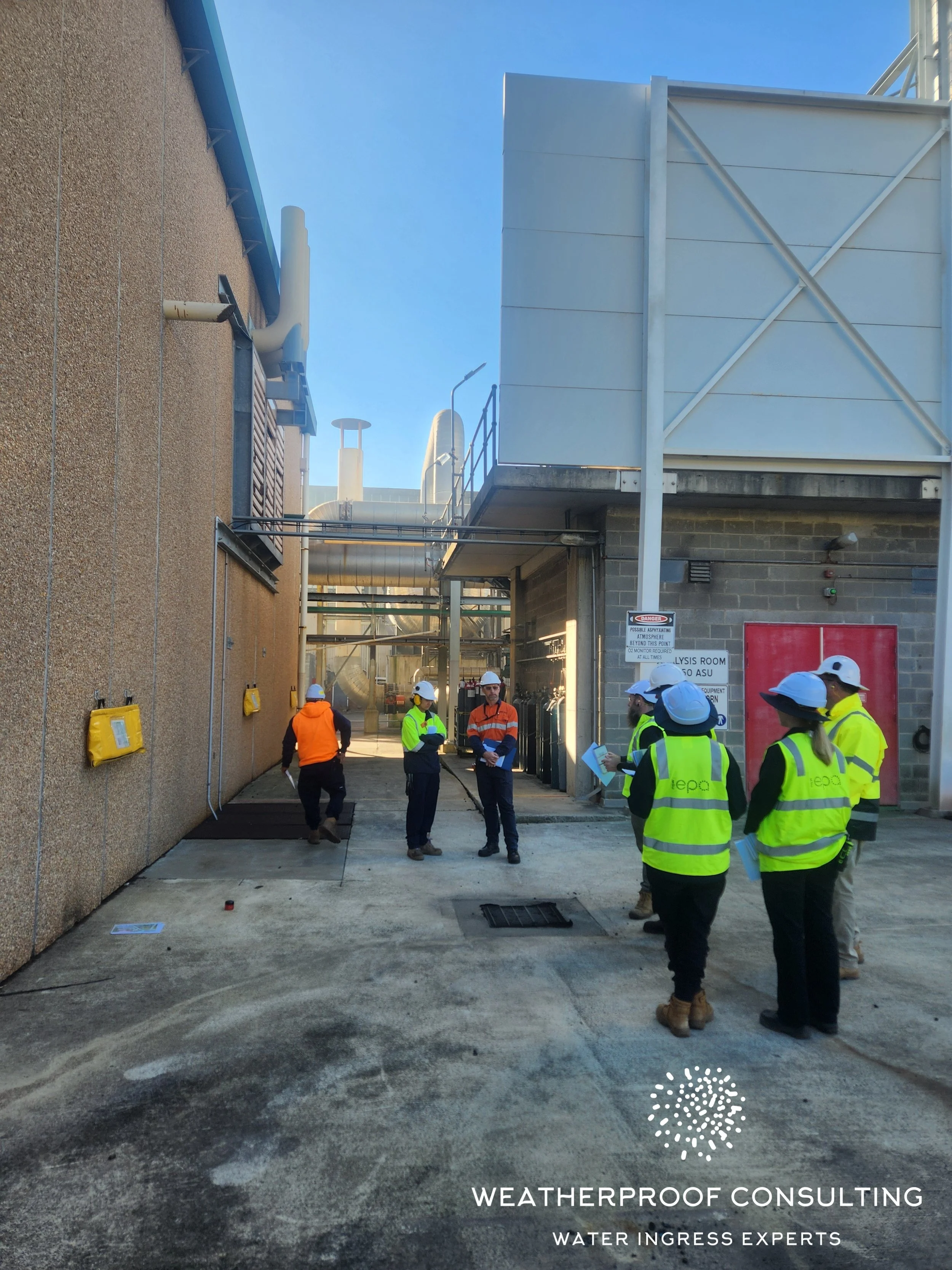 A group of workers wearing safety helmets and high-visibility vests stand and converse outside a building with industrial ventilation systems and ducts. There are warning signs related to weatherproofing and water ingress.
