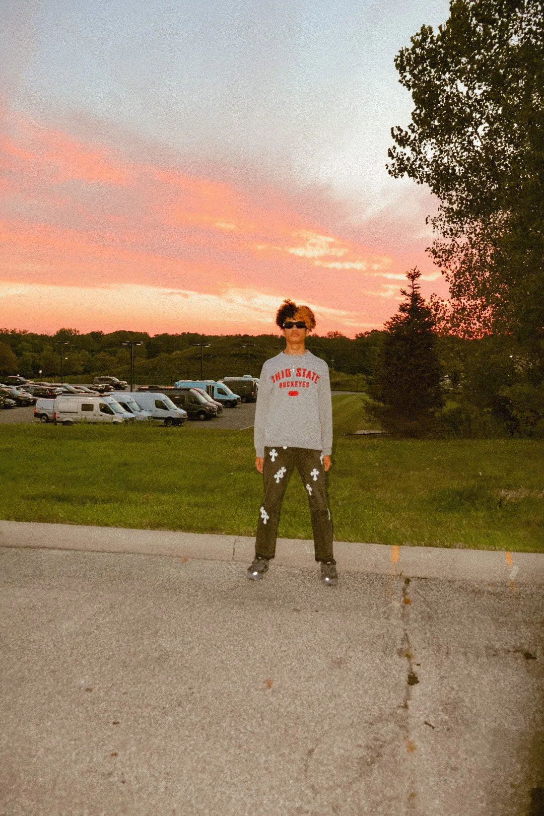 Person standing outdoors on pavement during sunset, wearing a gray Cleveland State sweatshirt, black pants with white graphics, gray shoes, and sunglasses, with a parking lot and trees in the background.