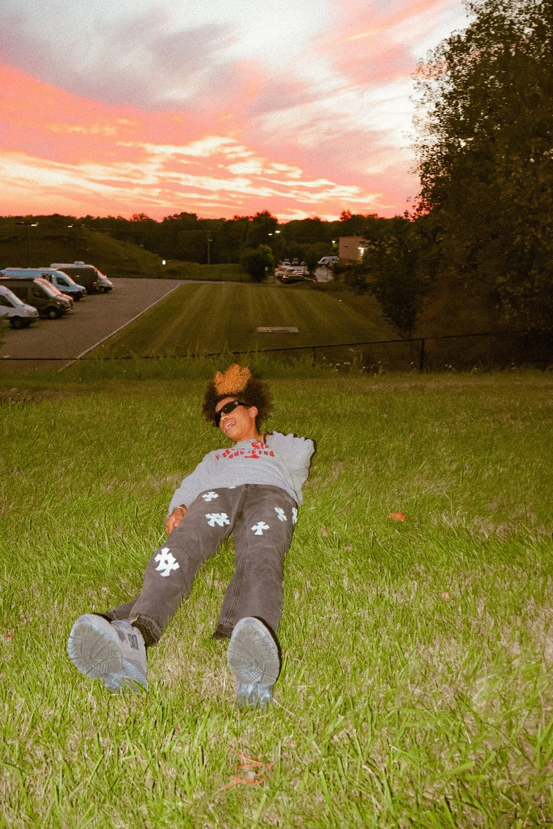 A person lying on a grassy hill at sunset, smiling and wearing sunglasses, with cars and trees in the background.