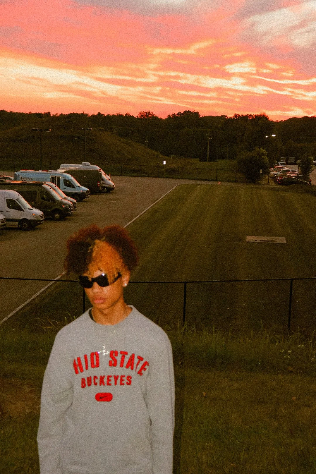 A young person with curly hair wearing sunglasses and a gray Ohio State Buckeyes sweatshirt, standing outdoors at sunset with a parking lot and grassy area behind them.