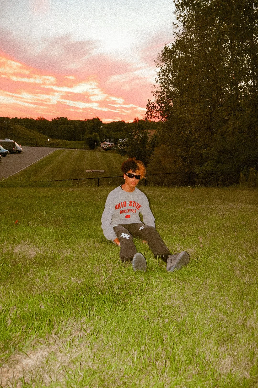 A person with curly hair and sunglasses sitting on a grassy hill during sunset, wearing a gray Ohio State Buckeyes sweatshirt and black pants.