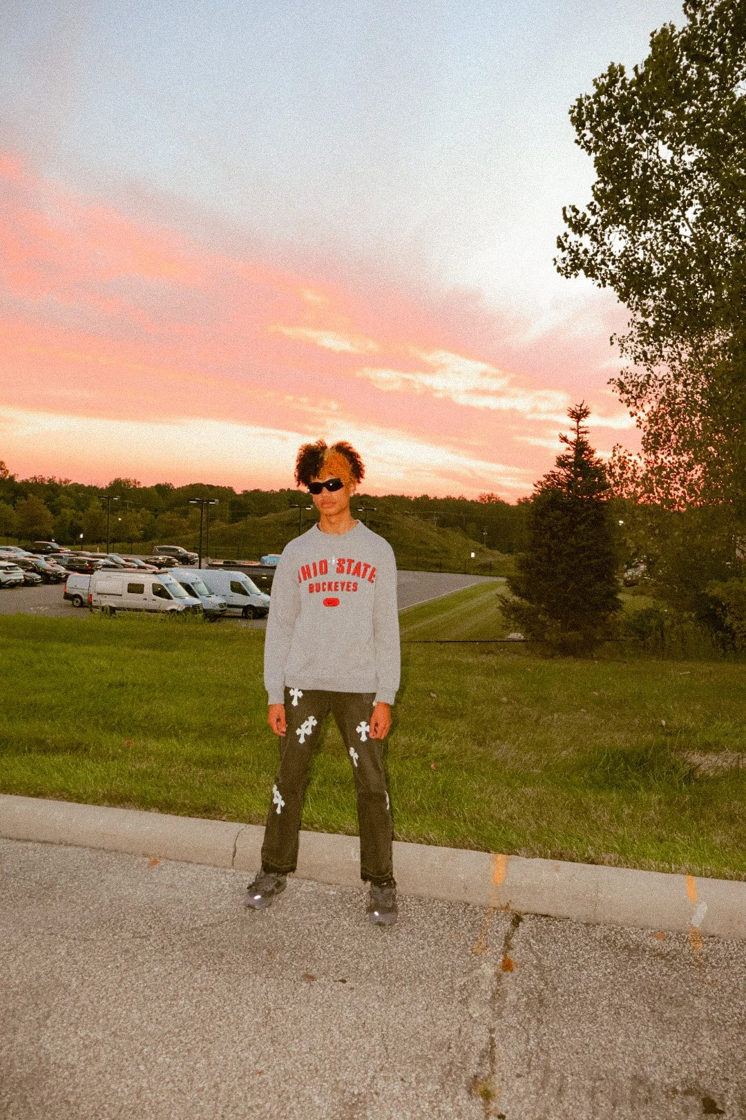 Person standing on curb in a parking lot during sunset, wearing sunglasses, a gray sweatshirt with Ohio State Buckeyes on it, black pants with white designs, rollerblades, with trees and parked cars in the background.