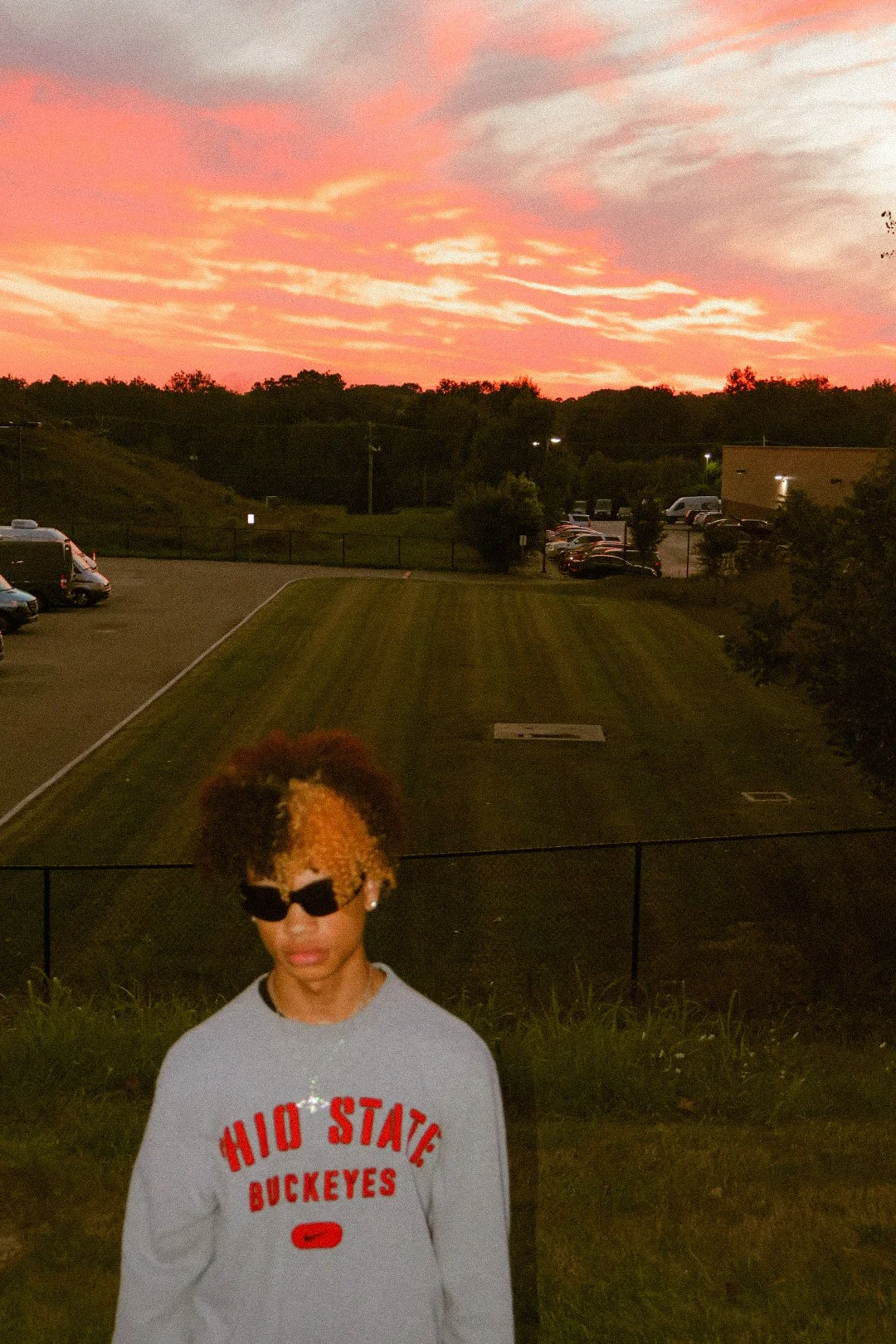 A person with sunglasses and curly hair, wearing an Ohio State Buckeyes sweatshirt, standing outdoors during sunset with pink and orange clouds in the sky, a grassy area, parked cars, and trees in the background.