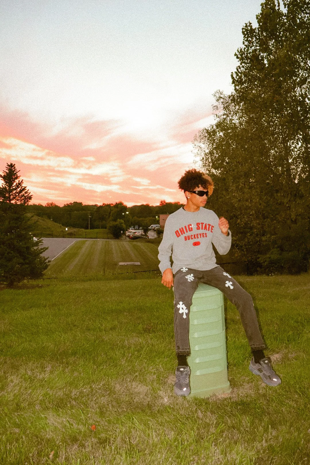 Young man sitting on a green sandbox outdoors during sunset, wearing a gray Ohio State Buckeyes sweatshirt, camouflage pants with white flower prints, sunglasses, and sneakers.