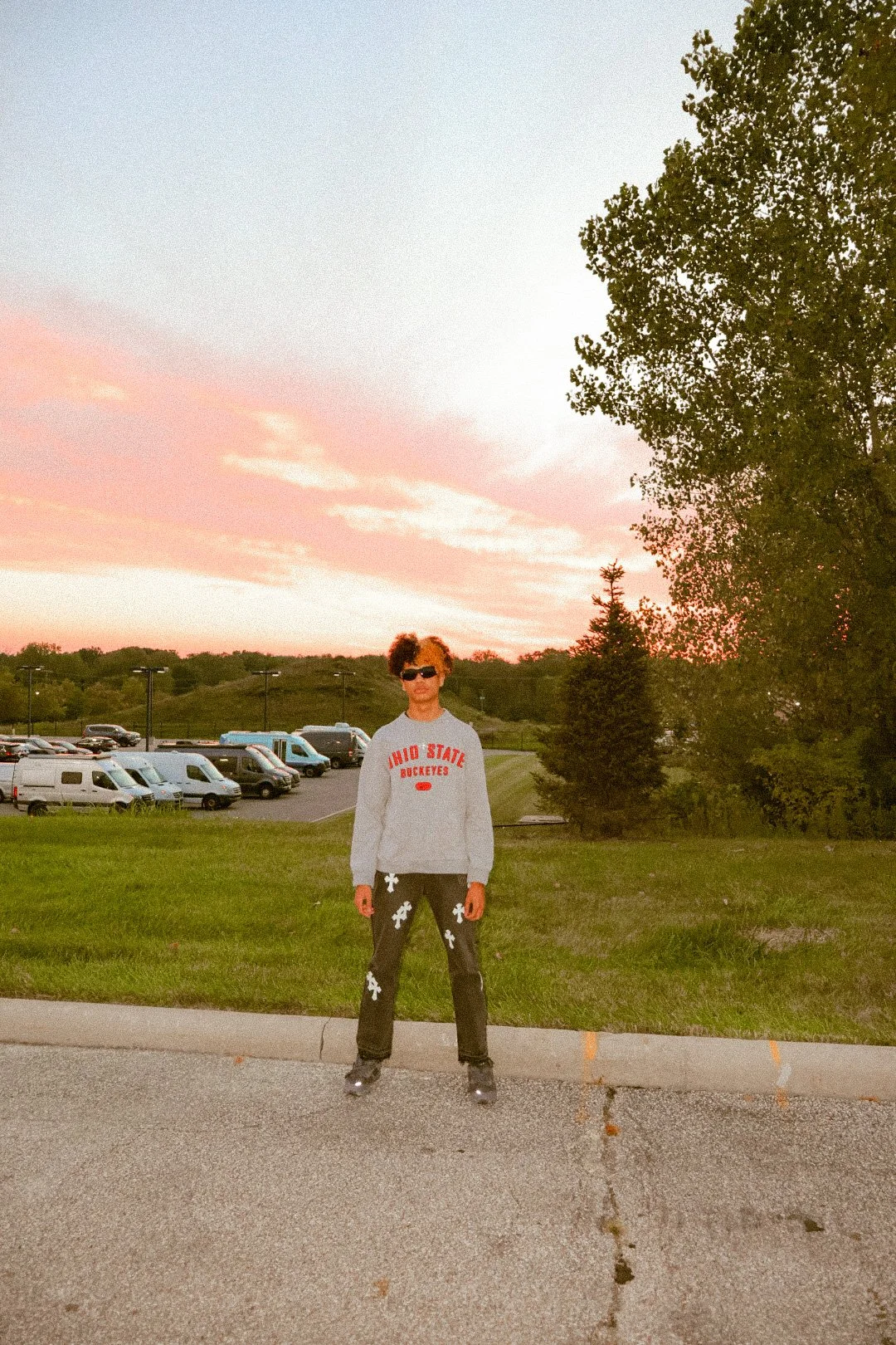 Person wearing sunglasses, Ohio State hoodie, black pants with white designs, and roller skates standing on a sidewalk during sunset with trees and parked cars in the background.