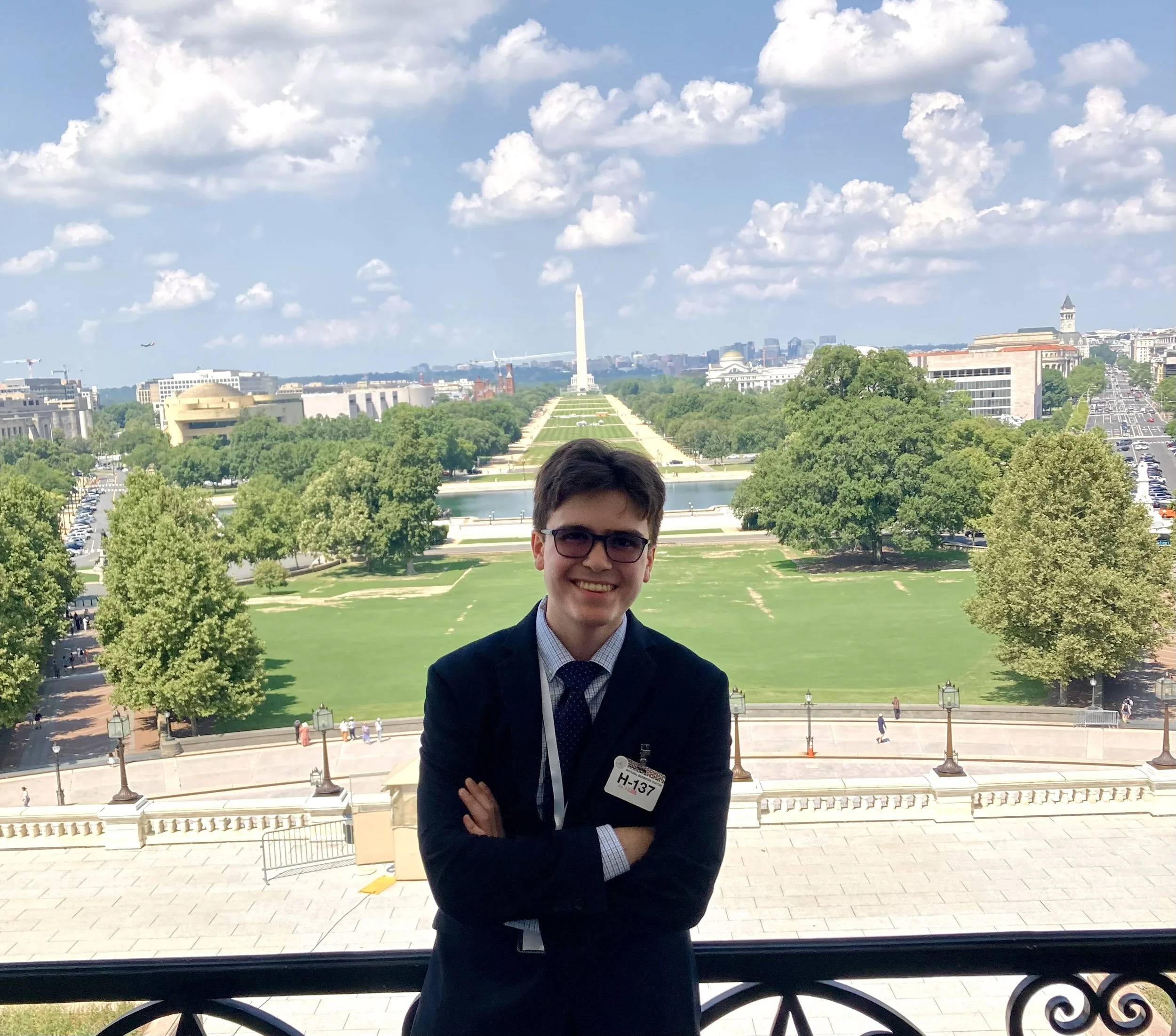 A young man with glasses and a suit stands with crossed arms in front of a railing, with the Washington Monument and the National Mall in the background on a sunny day with scattered clouds.