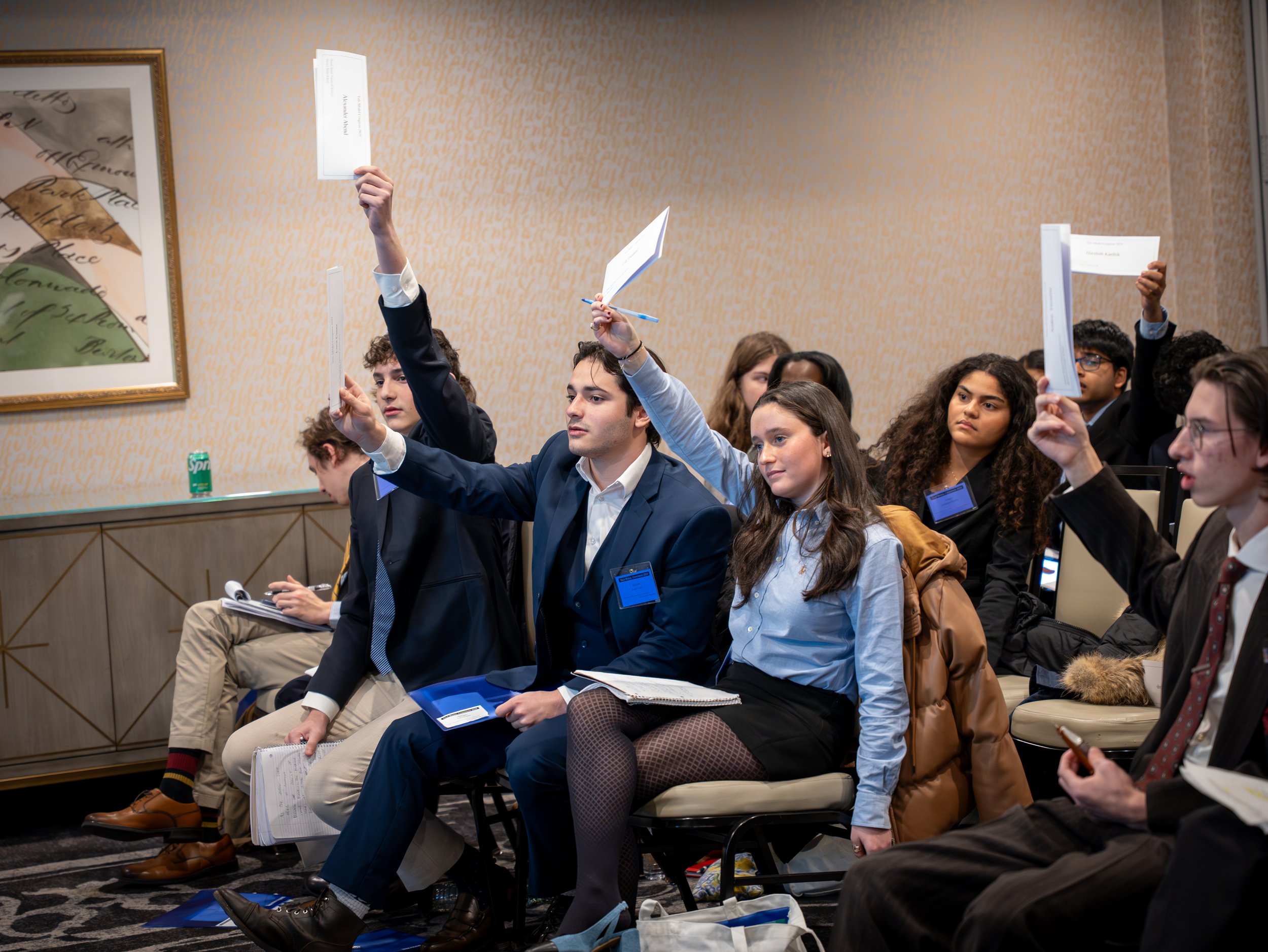 Young adults in formal attire attending a conference or seminar, with some raising their hands to ask questions, seated in a room with beige wallpaper, framed art on the wall, and note-taking materials.