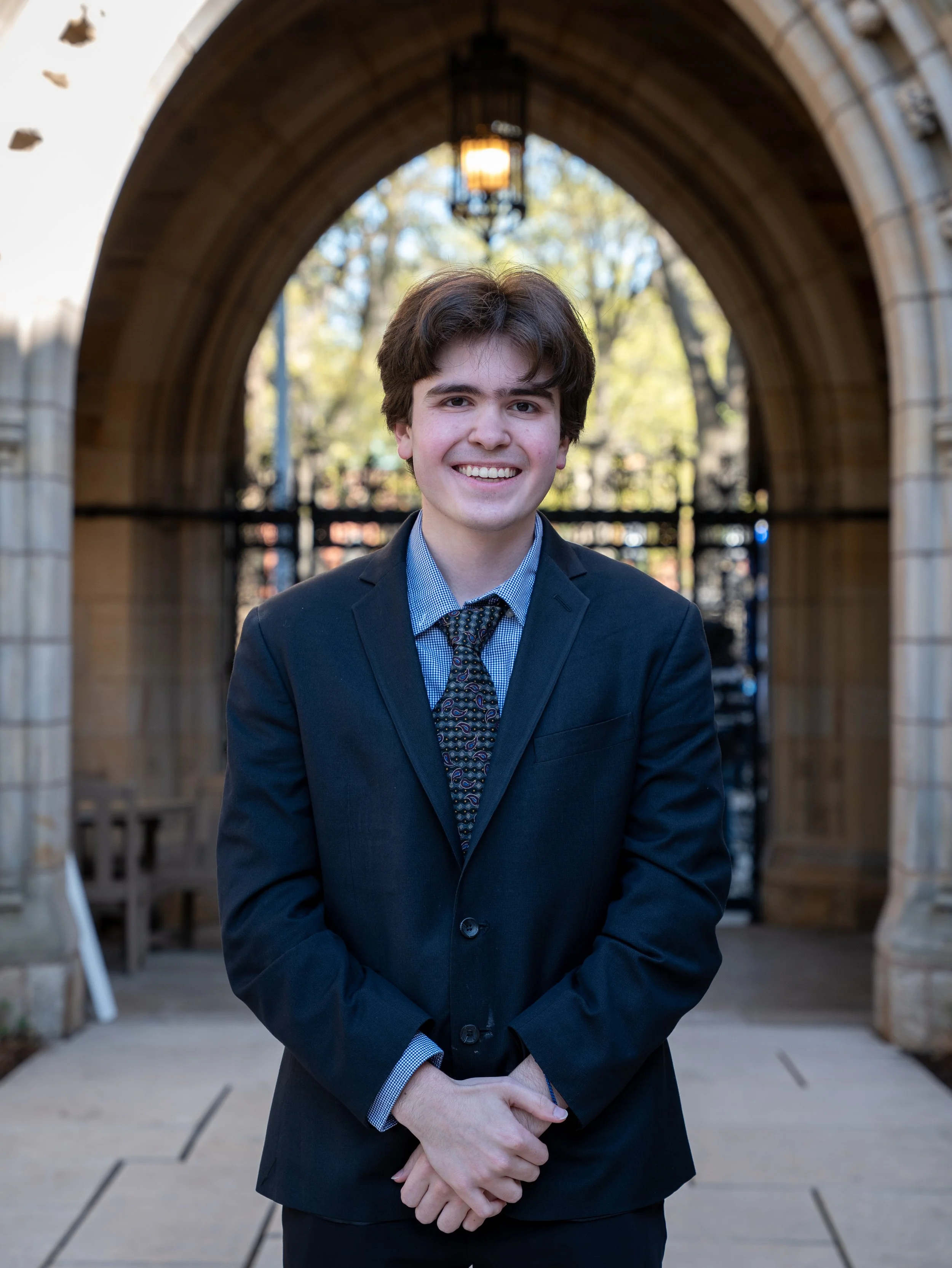 A young man in a dark suit smiling, standing under a stone archway with trees in the background.