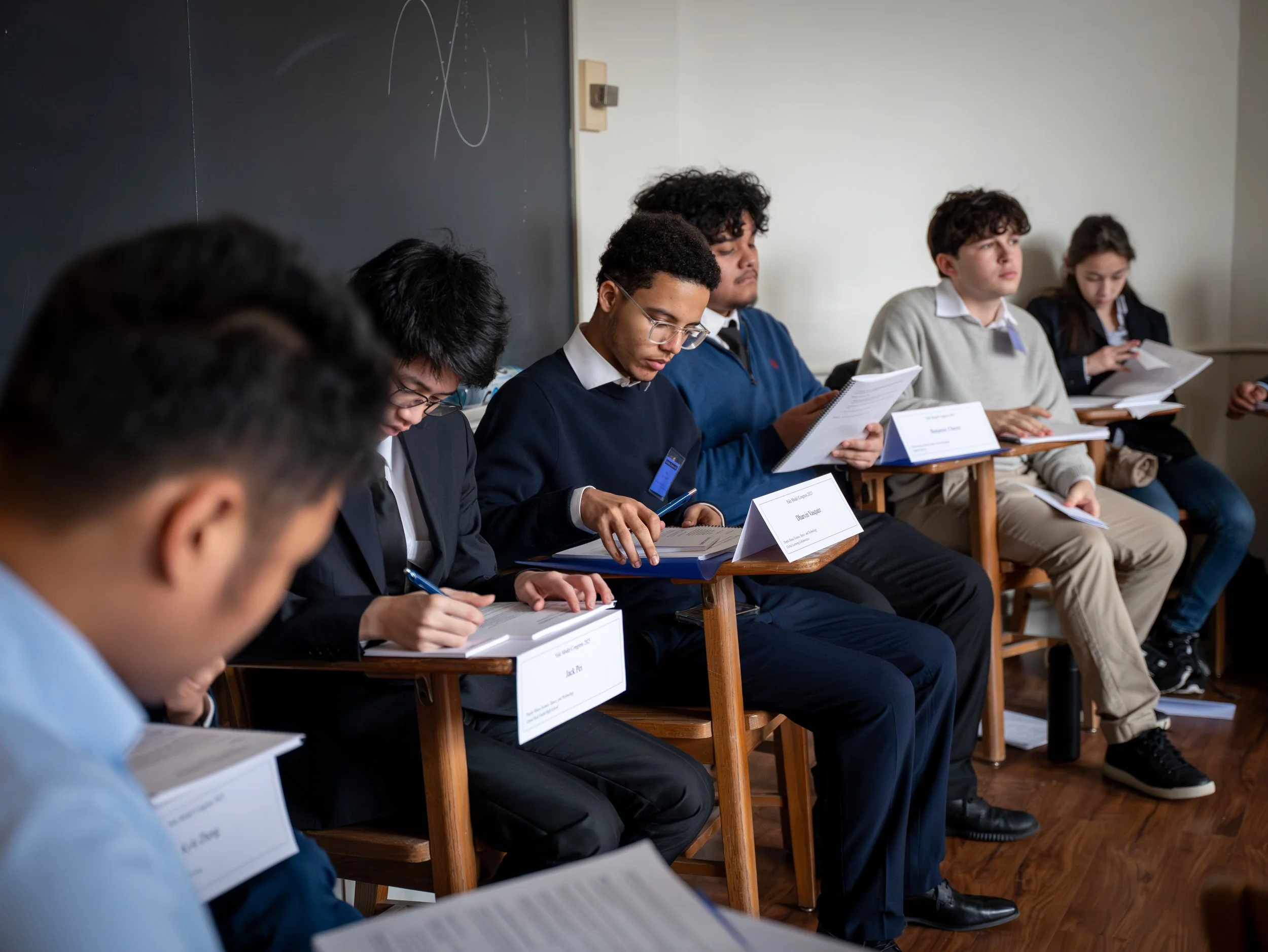 A group of students sitting on wooden chairs in a classroom, reading papers and taking notes, with a blackboard behind them.