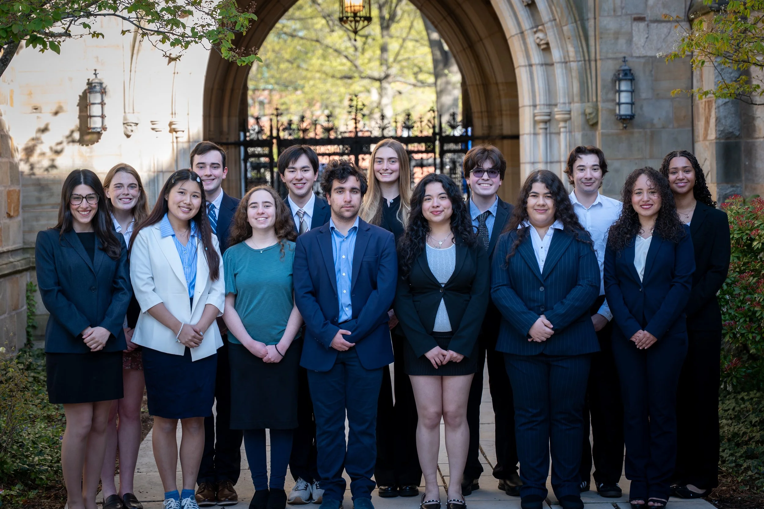 A diverse group of students dressed in formal attire, smiling, standing outside a historic stone building with an arched entrance and green trees in the background.