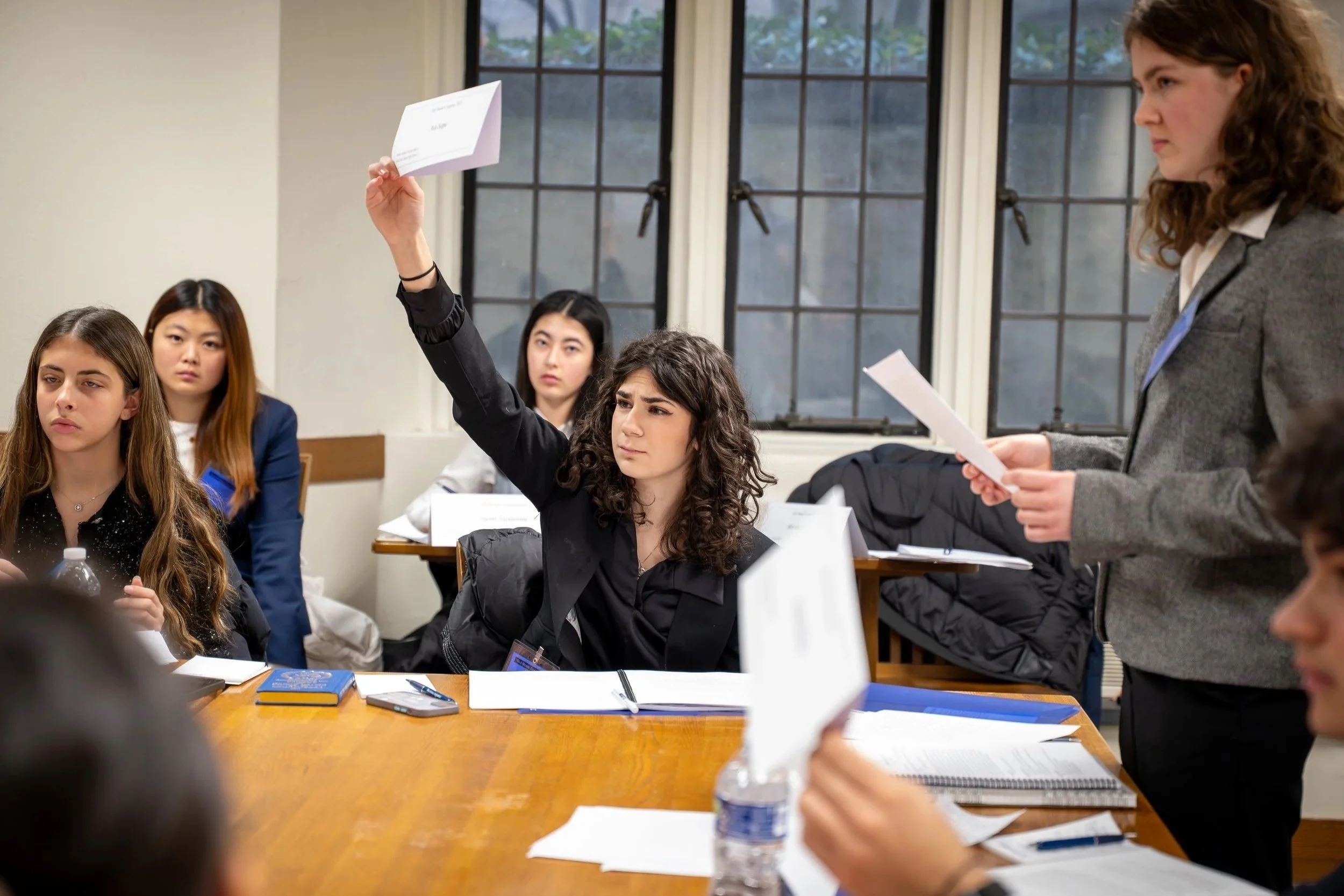 Young woman raising her hand in a classroom setting with other students listening and a woman standing and speaking.