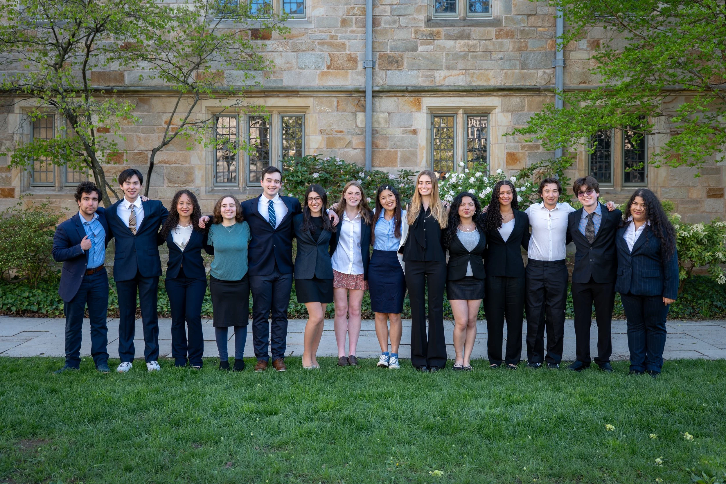 Group of diverse young adults standing outdoors on a grassy area, smiling and posing for a photo, with an old stone building and trees in the background.