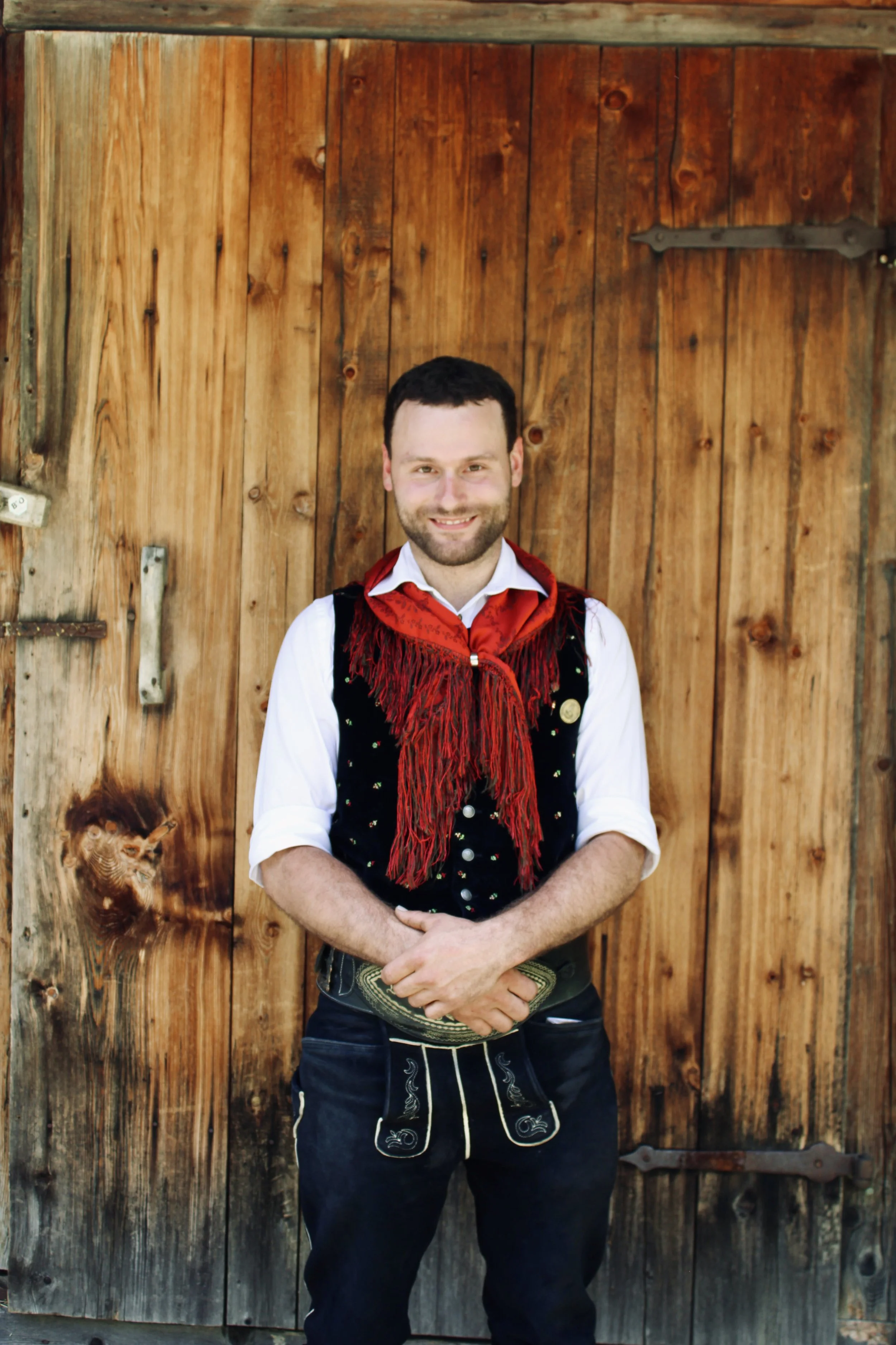 Ein lächelnder Mann mit dunklen, lockigen Haaren und Bart steht vor einem grauen Vorhang, trägt eine weiße Hemd, eine schwarze Weste mit roten Punkten, eine rot-braune Fransen-Schleife um den Hals und hält ein Smartphone in beiden Händen.