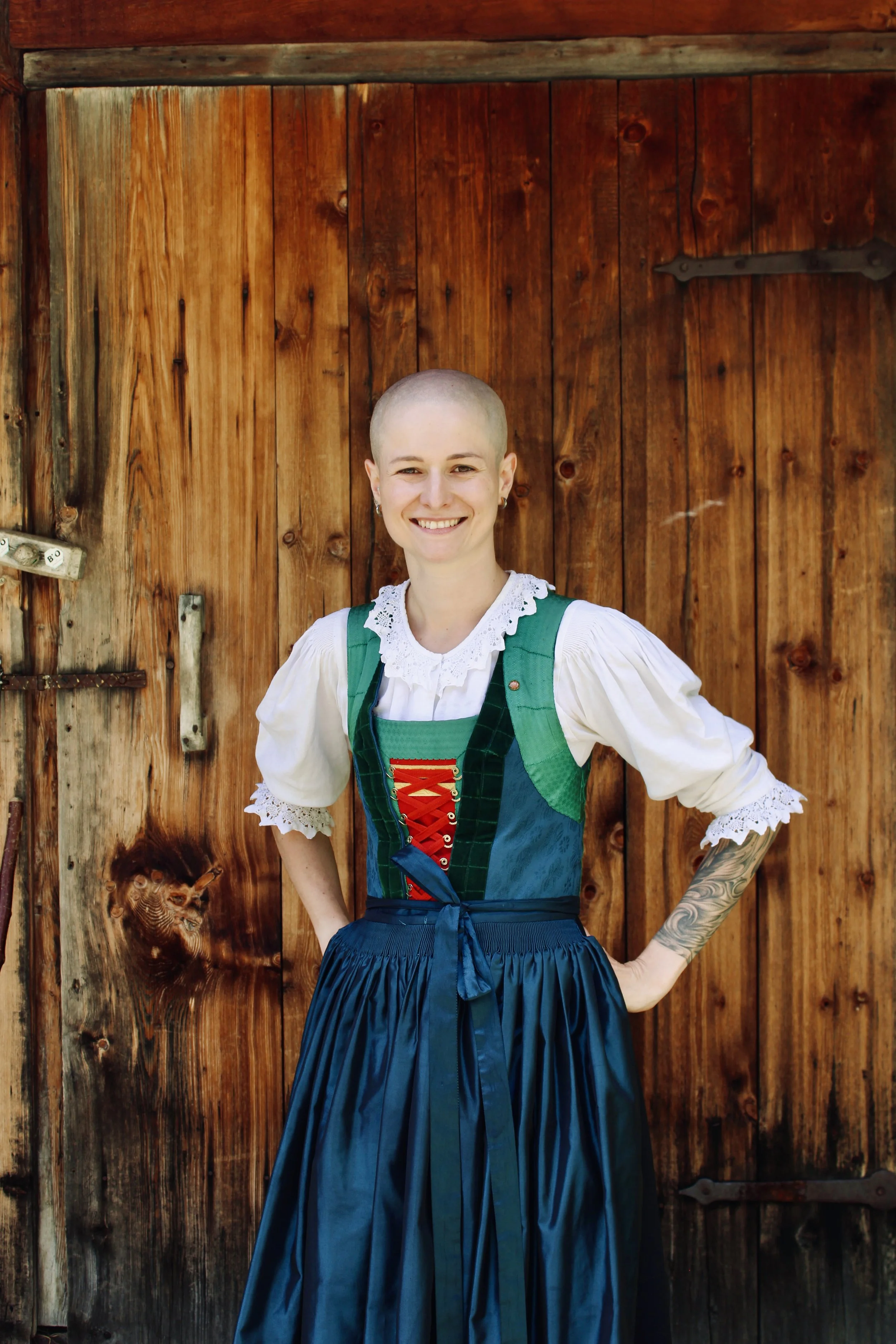 Junge Frau in traditioneller Tracht, die vor einem Haus und grüner Landschaft in den Bergen steht, bei sonnigem Wetter.