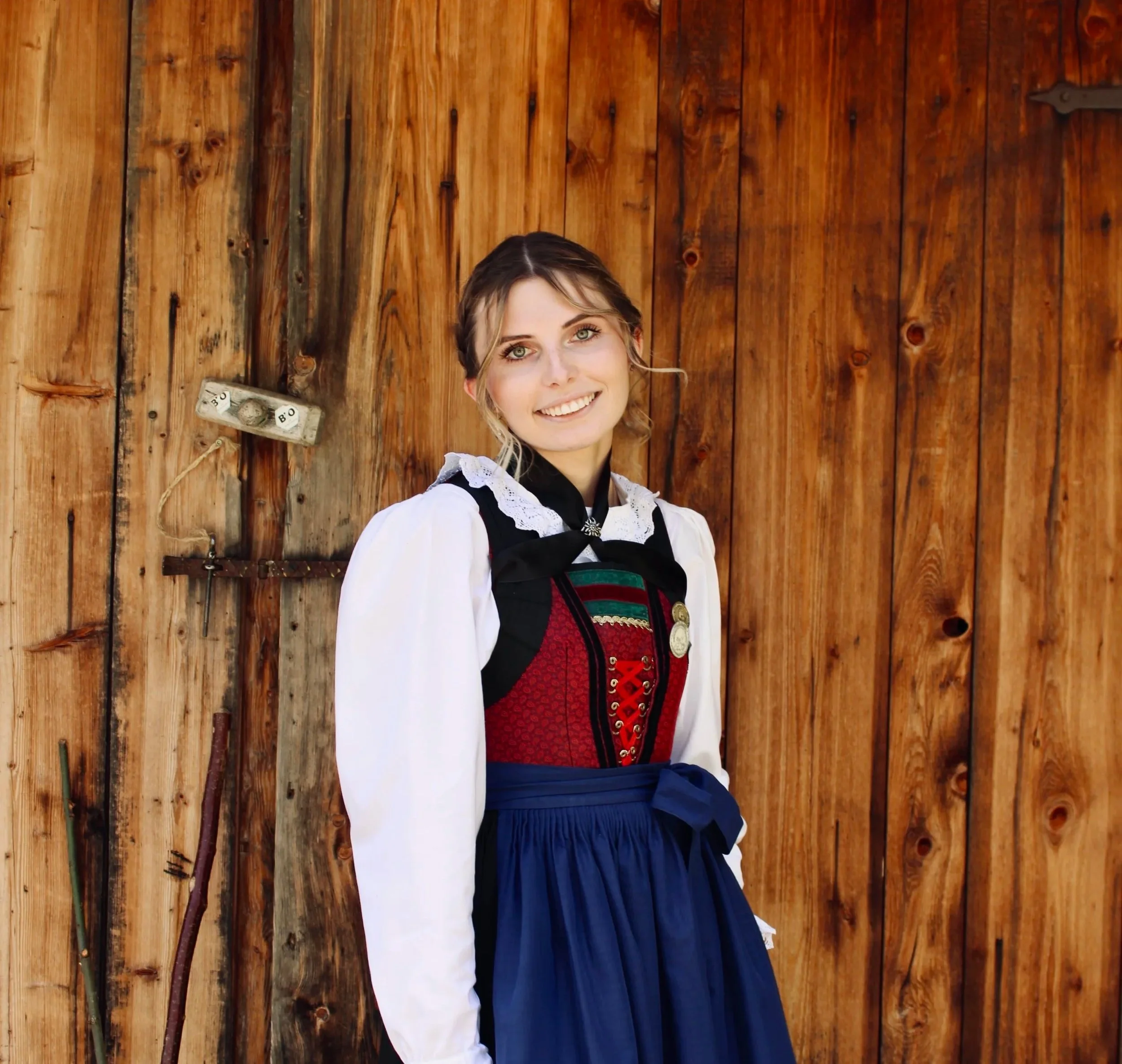 Junge Frau in traditioneller Kleidung im Freien vor einem Haus mit Bergen im Hintergrund.