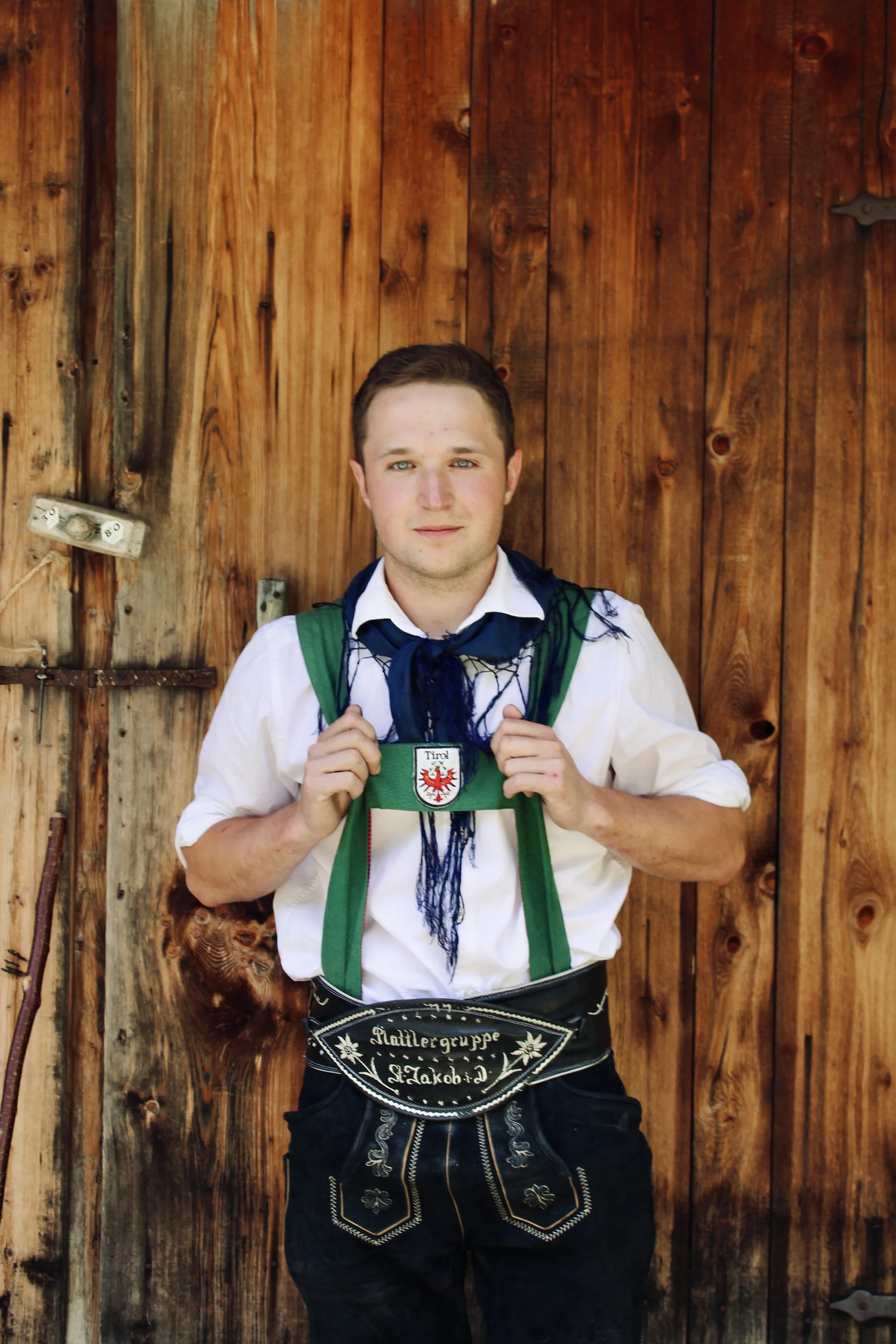 Junge Mann in weißem Hemd mit grünem und schwarzem Schal, der eine Fahne mit Wappen trägt, vor grauen Vorhängen.