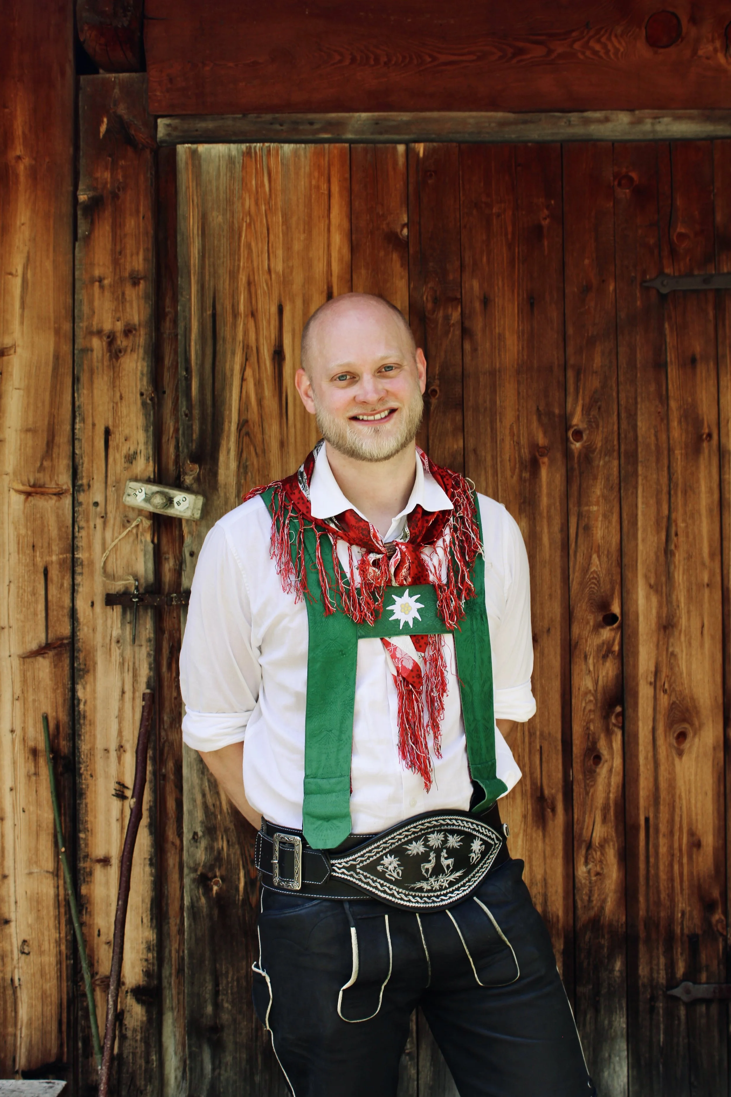 Ein Mann in traditioneller Tracht mit weißen Hemd, roten Schleifern, grüner Hacke, schwarze Lederhose und schwarzer Gürteltasche, vor grauem Vorhang.