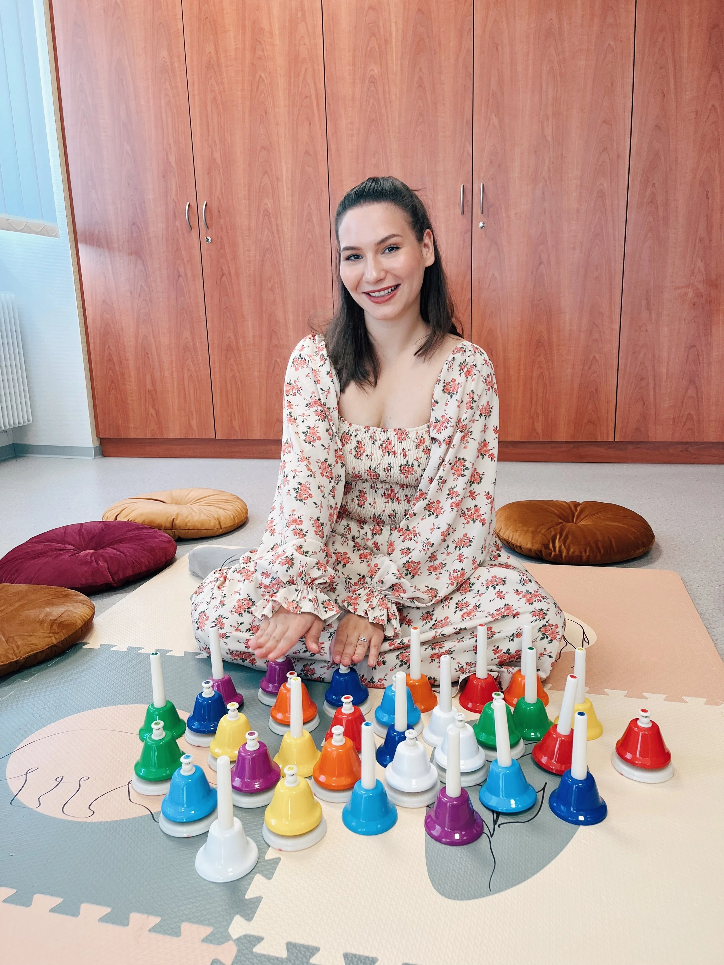 A woman sitting on the floor of a room with wooden cabinets behind her, smiling, wearing a floral dress, surrounded by colorful bells on the floor in front of her.