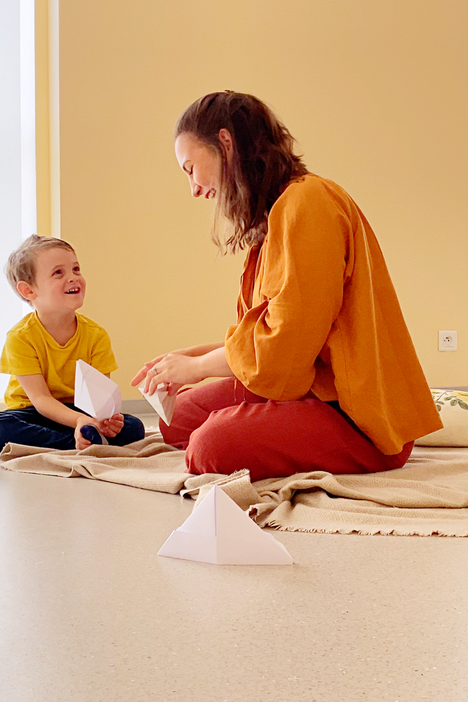 A woman and a young boy sitting on a blanket on the floor, playing with paper boats, smiling and laughing together in a cozy room.