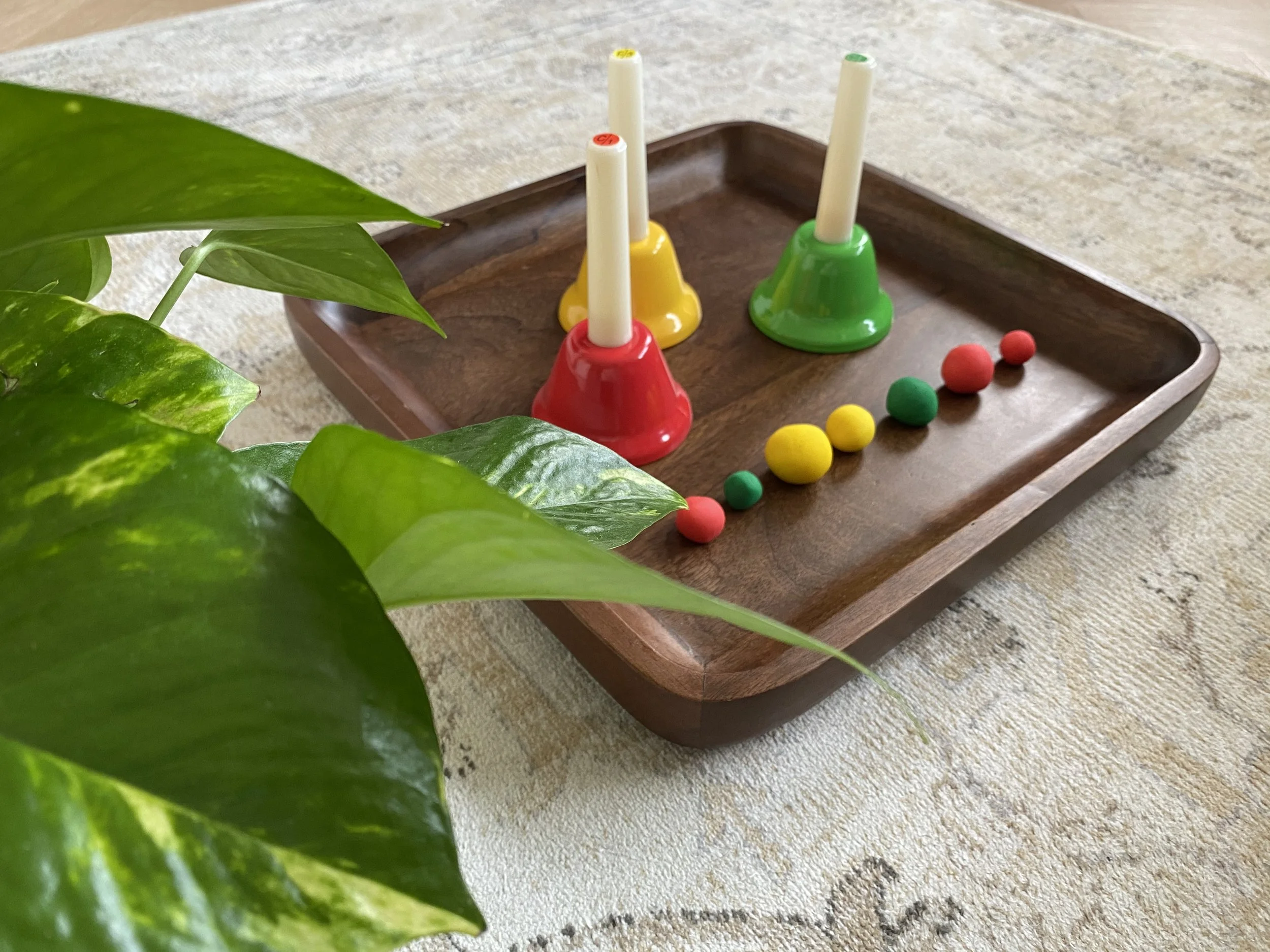 Colorful children's game set on a wooden tray, with three bell-shaped bases in red, yellow, and green, each with a white stick, and small colorful balls in yellow, red, green, and pink.