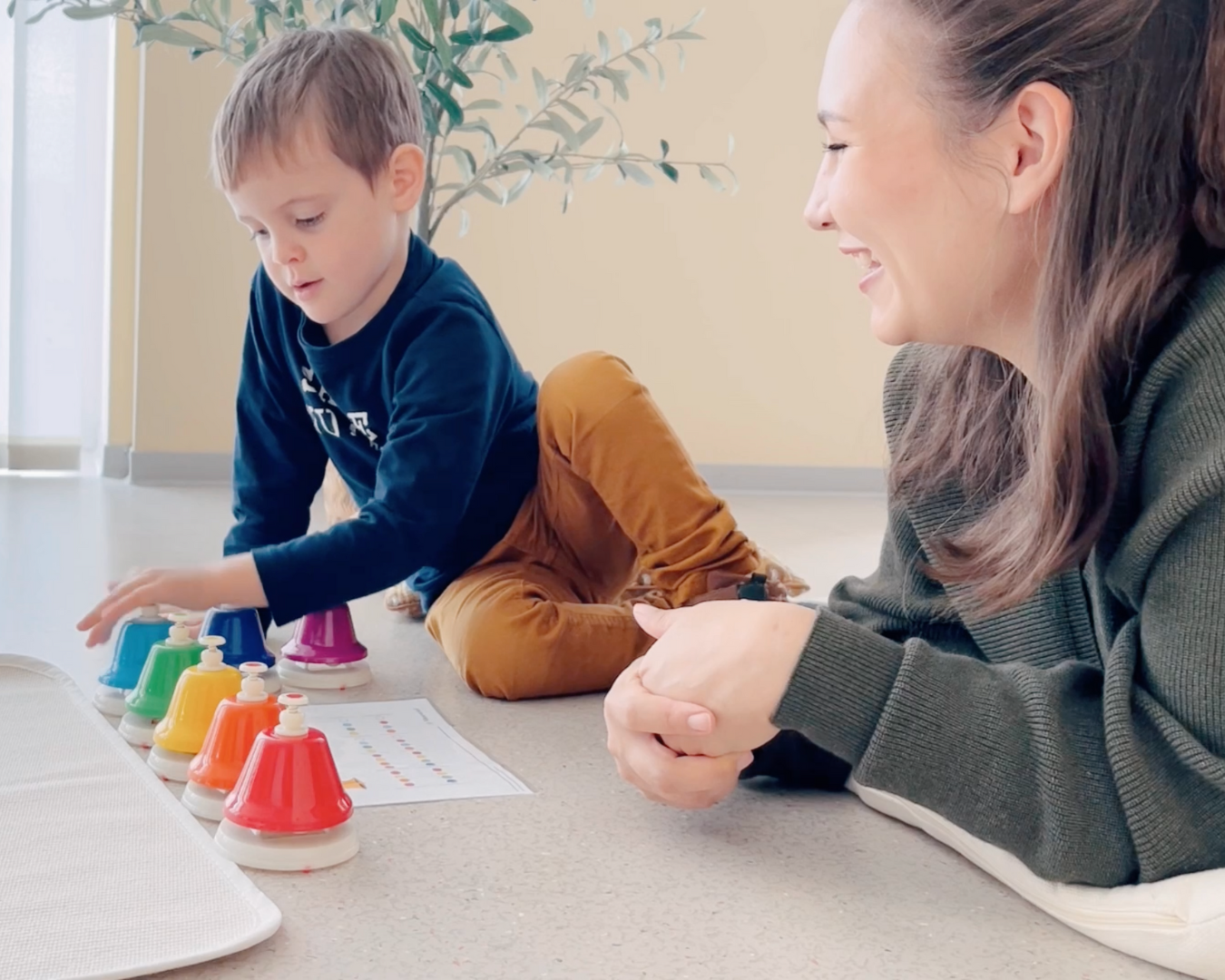 A young child and a play based music teacher are playing a game with colorful bells, smiling and engaged in the activity.