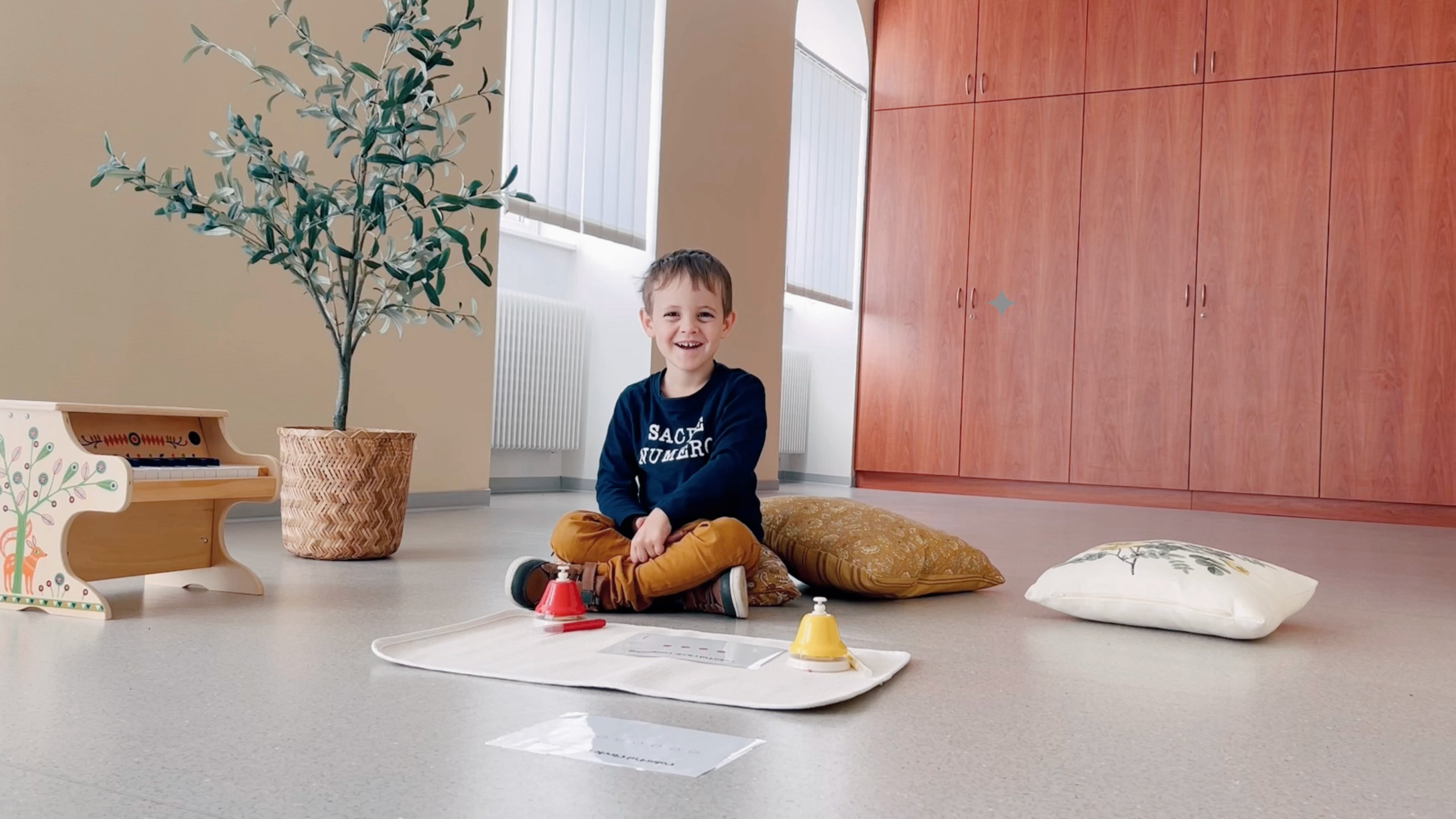 A young boy sitting cross-legged on the floor, smiling, with cushions behind him, a toy piano decorated with a deer and trees, a large potted plant, and a wooden cabinet in the background.