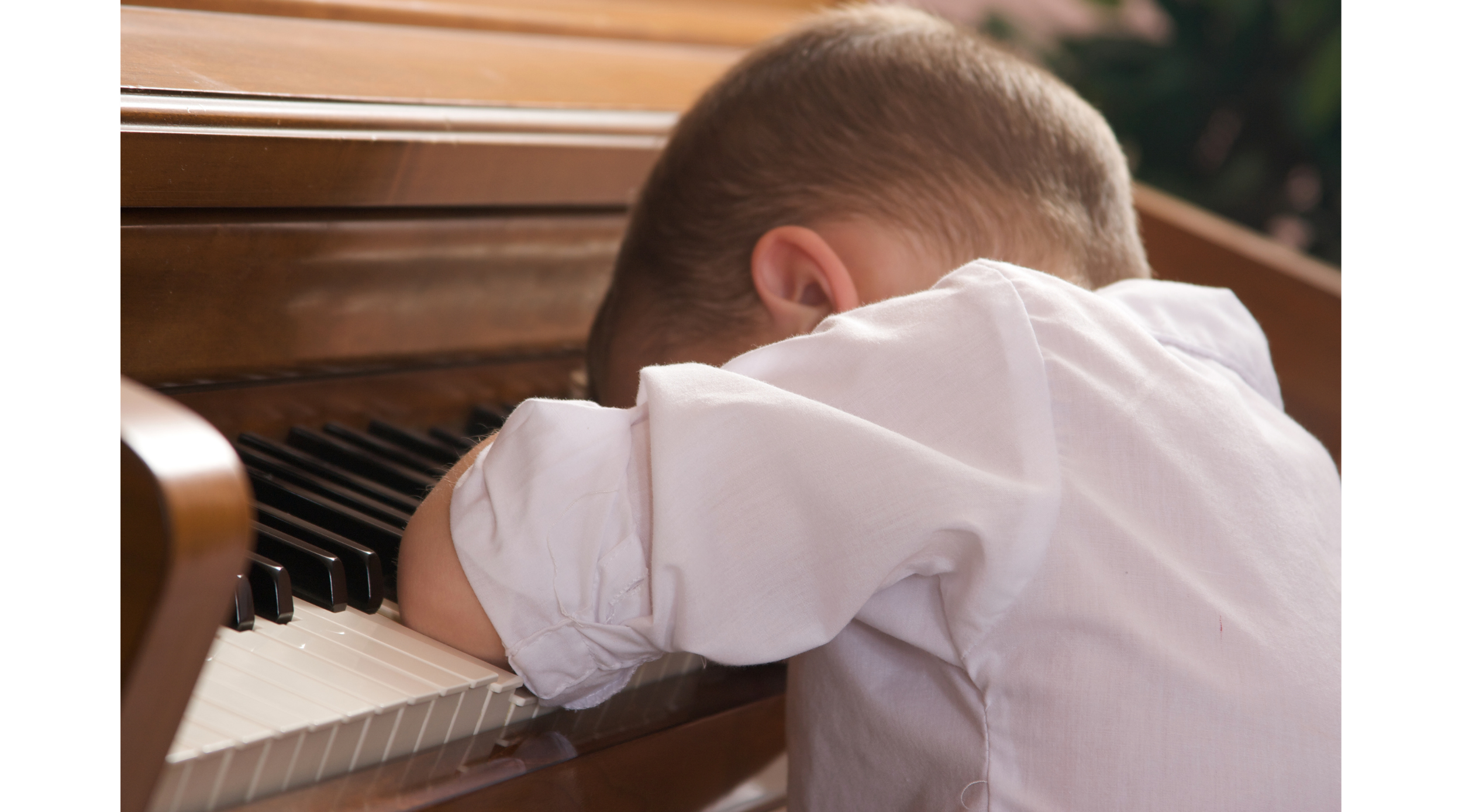 A young boy with short hair wearing a white shirt, resting his head and arms on a piano keyboard with his eyes closed.