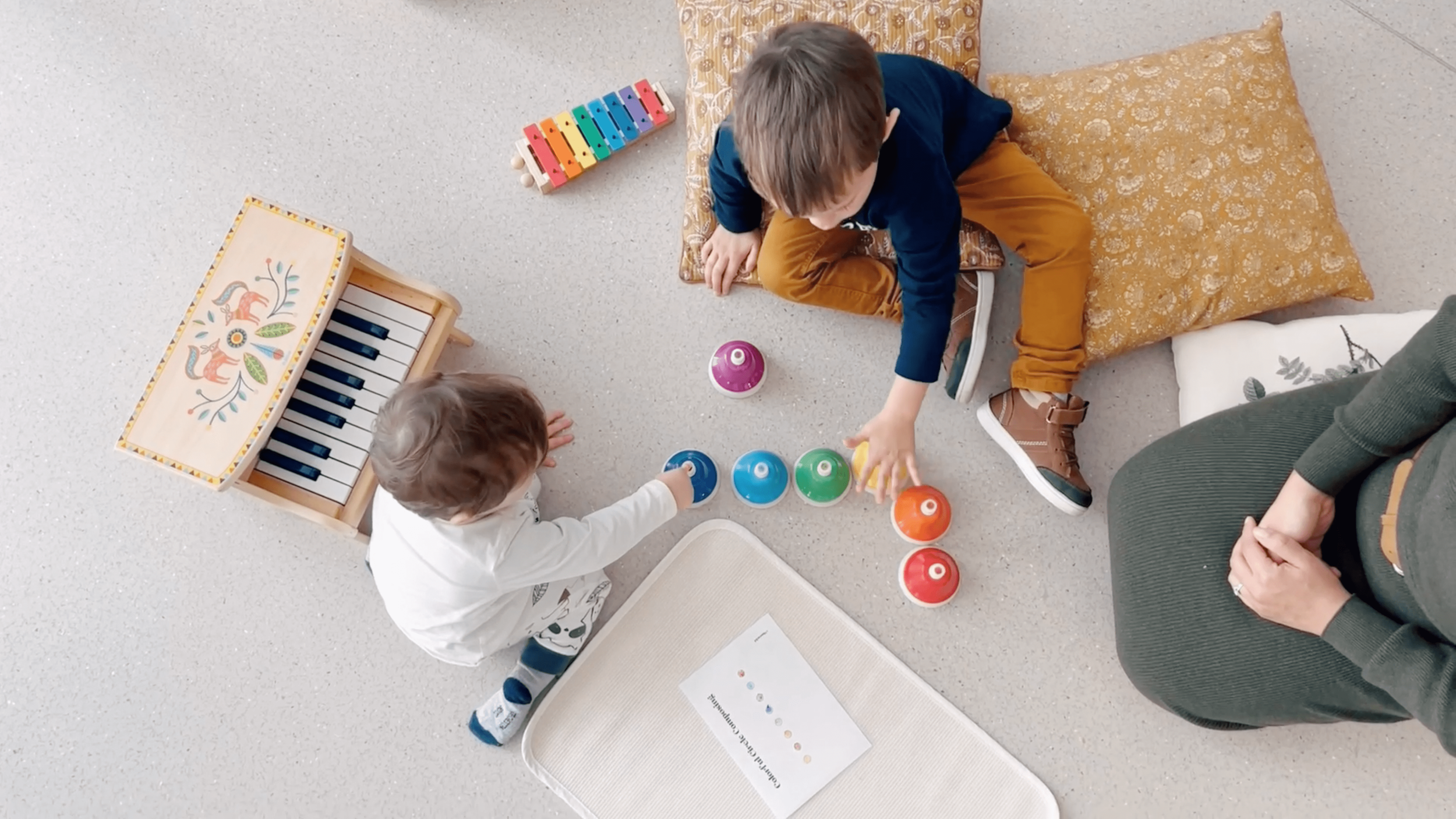 Two young boys playing with colorful game bowls on a light-colored floor, surrounded by some floor cushions and a small decorative wooden piano with painted animal designs. An adult woman is sitting nearby, observing the children.