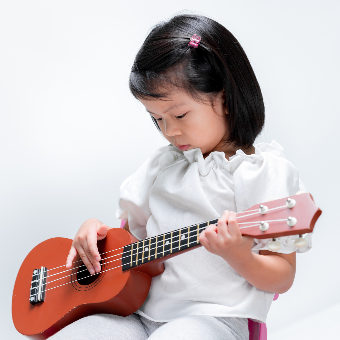 Young girl playing a small pink ukulele, looking down at the instrument, against a plain white background.