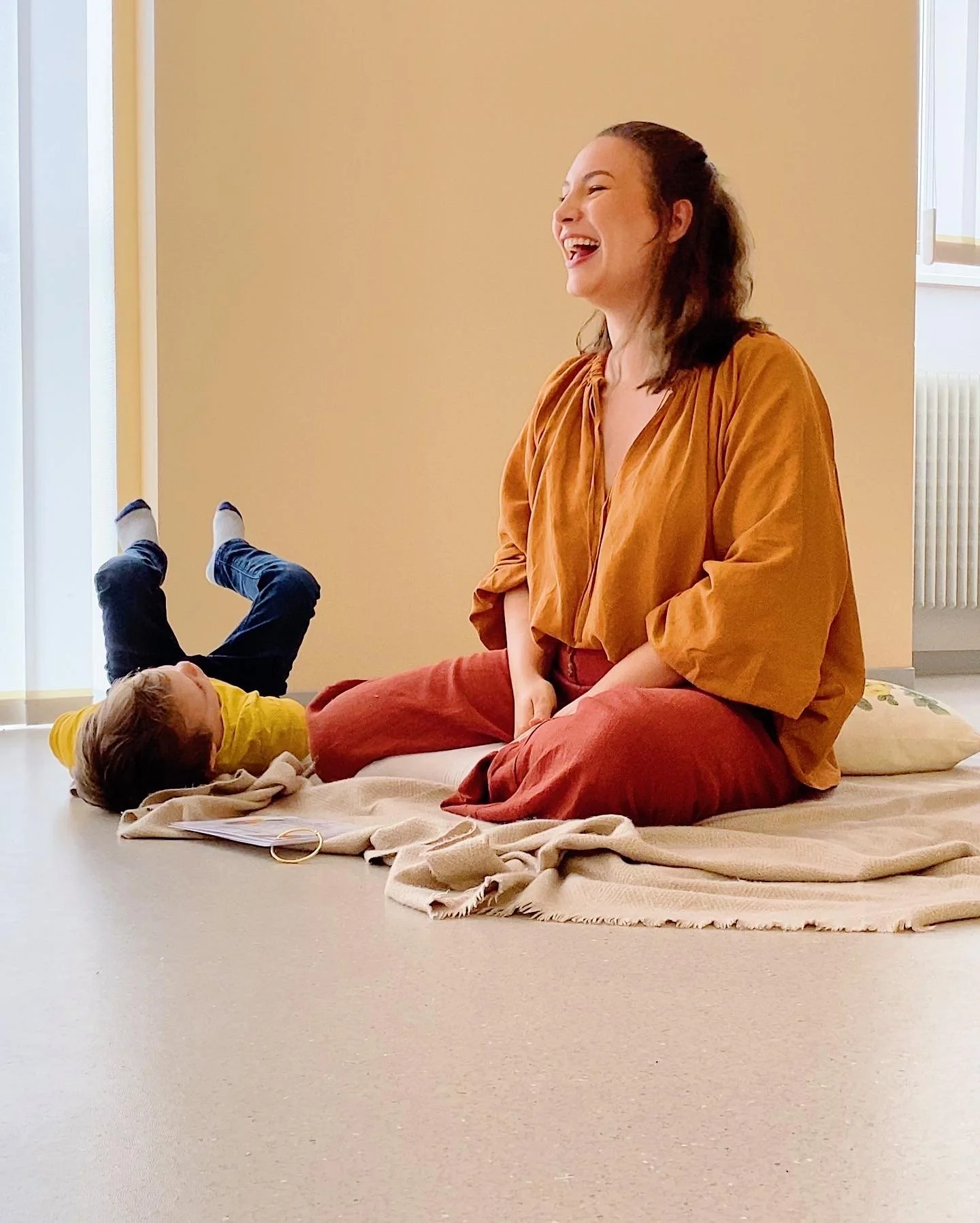 A woman sitting on a blanket on the floor, laughing, with a young child lying next to her, playing or resting.