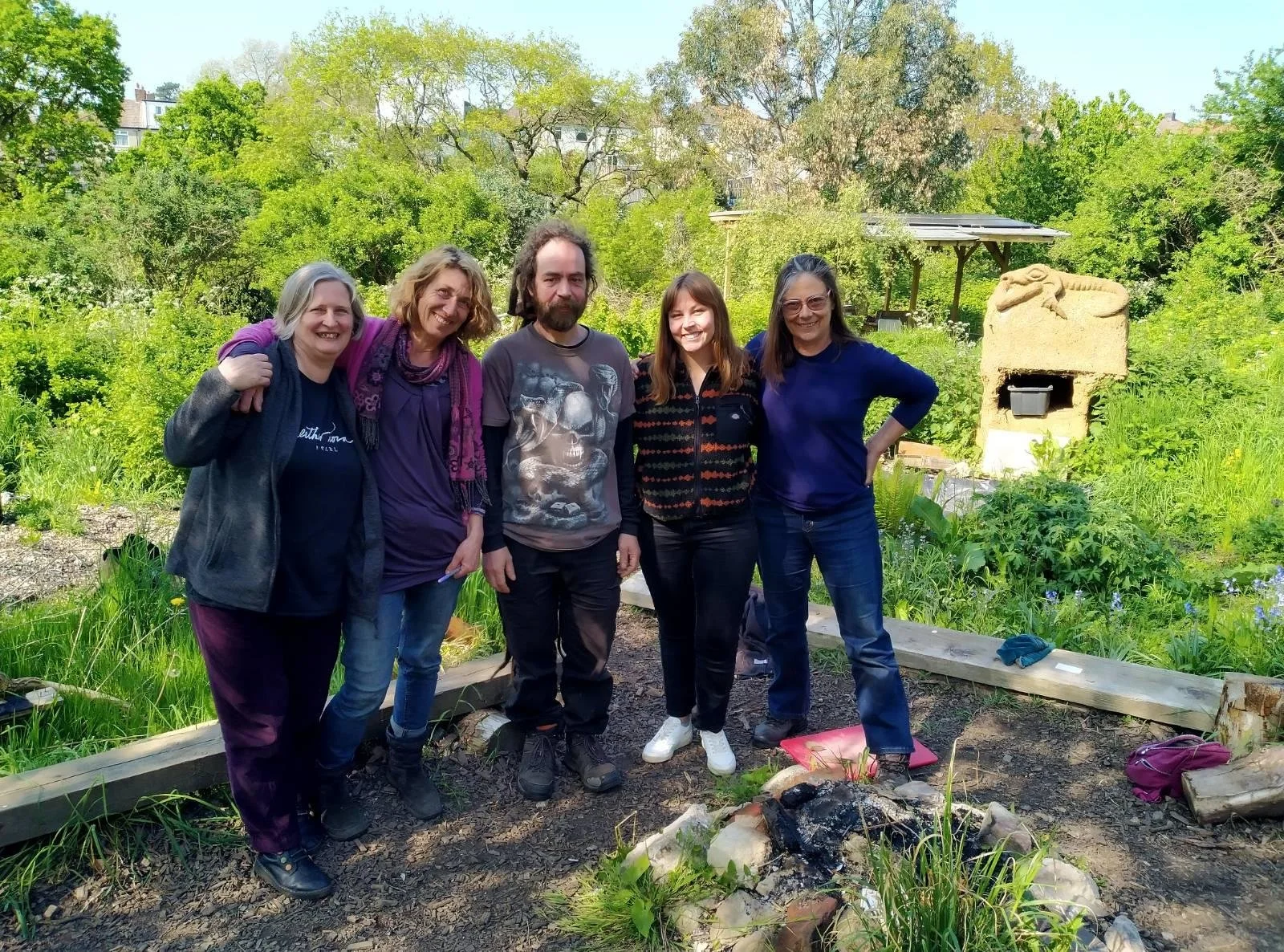 5 people stand between an unlit campfire and some wooden planks laid on the floor like benches. Some of them have their arms around each other, all of them are smiling.