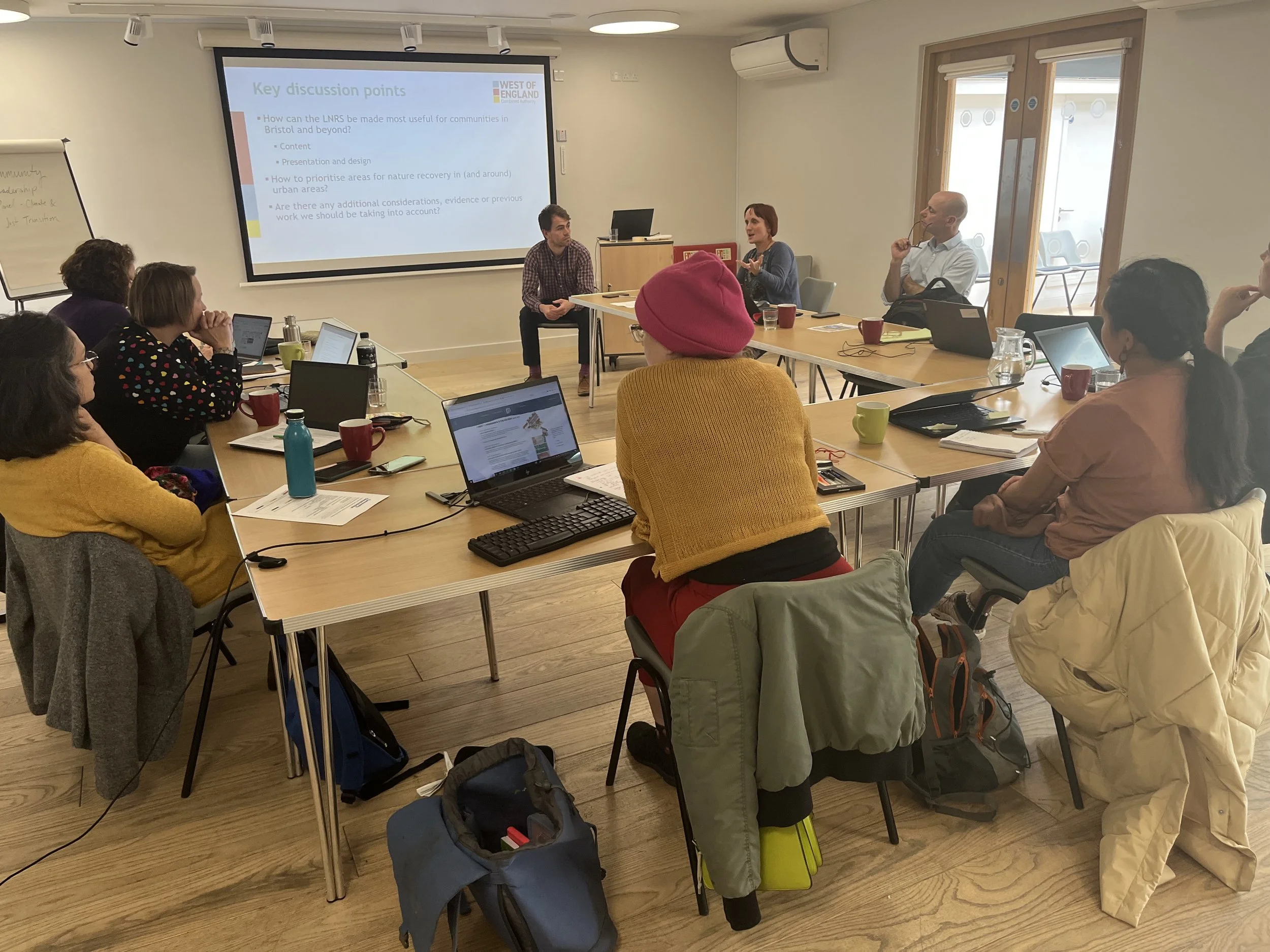 Two people are presenting a powerpoint. Another seven people sit around a table in a horseshoe shape, watching the presentation.