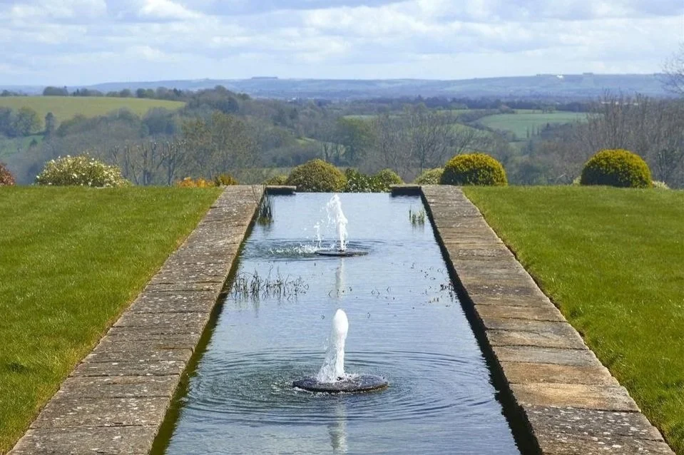 A fountain and pool of water extends towards green rolling fields in the distance. 
