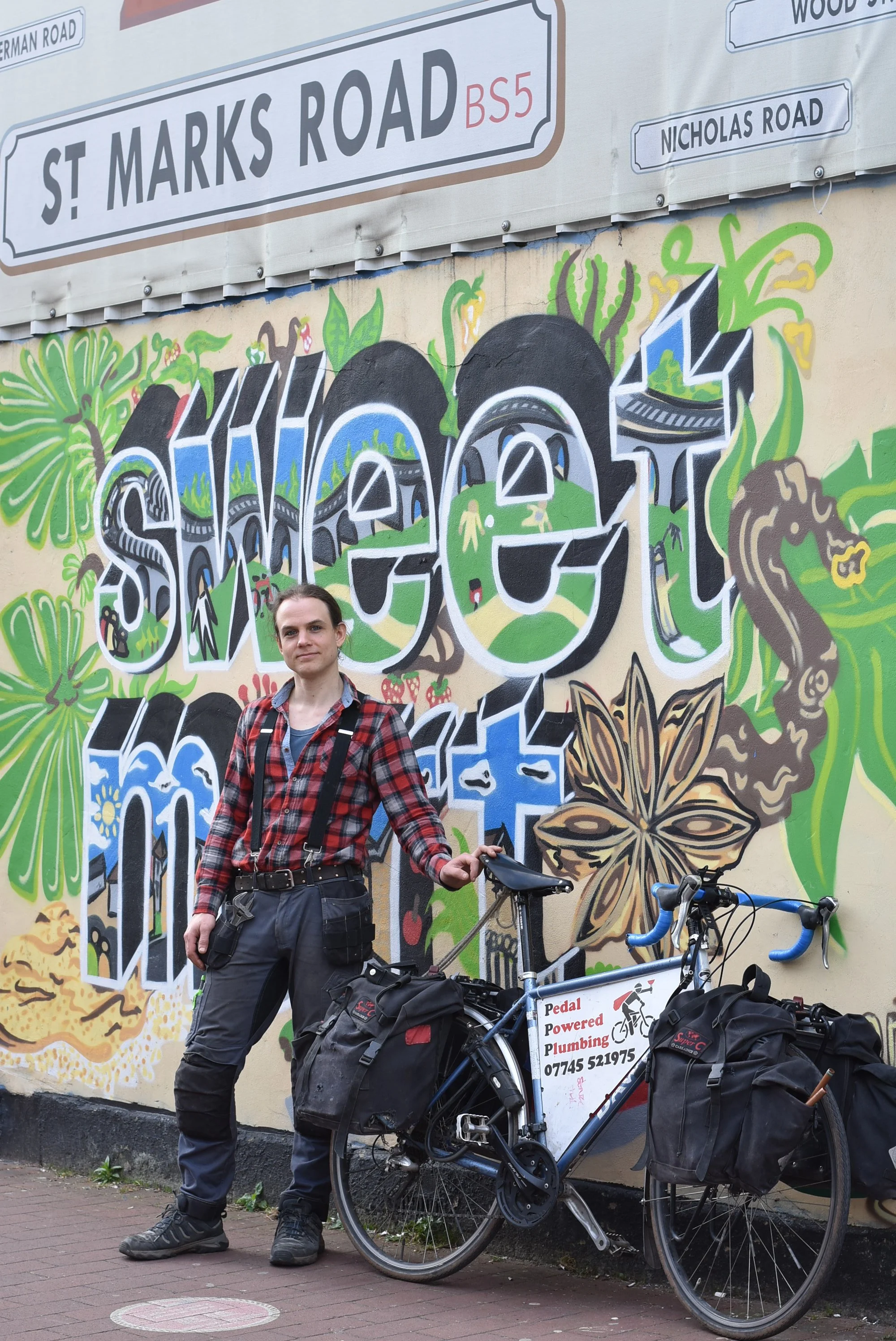 A plumber stands next to their push bike in front of street art reading 'sweet mart.'