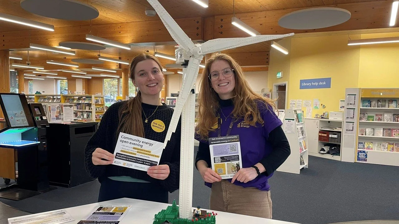 Two employees from the Centre for Sustainable Energy hold flyers and smile at the camera, behind a 3ft Lego wind turbine.