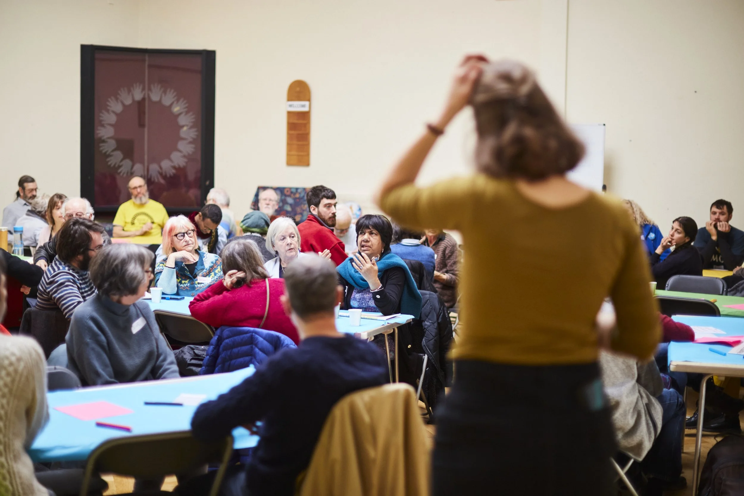 A room full of people sit around tables, with about 6 people per table.  Emilia is in the foreground with her back to the camera. 