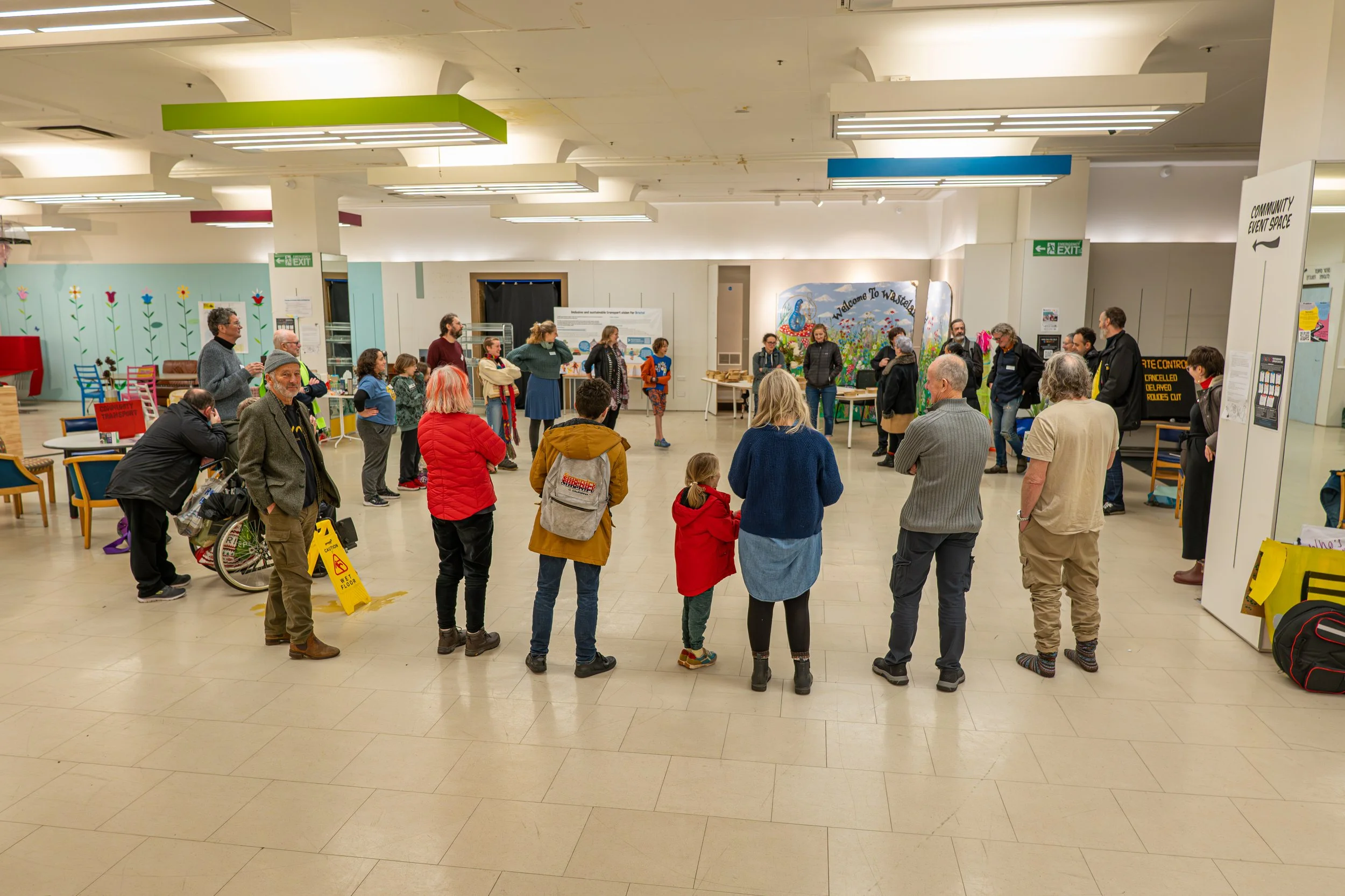 About 30 people stand in a circle in a large room. The room has white tiling floor.