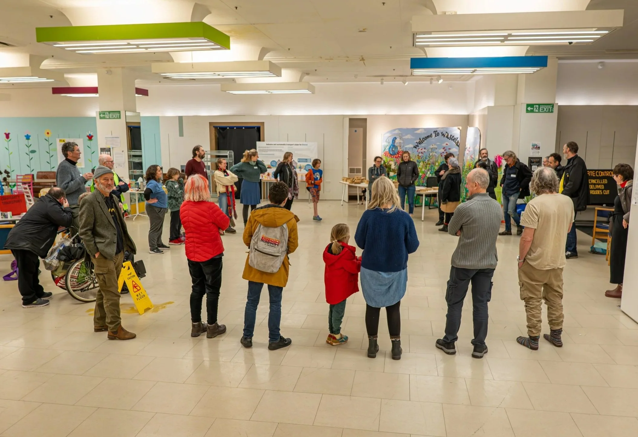 About 30 people stand in a circle in a large room. The room has white tiling floor.