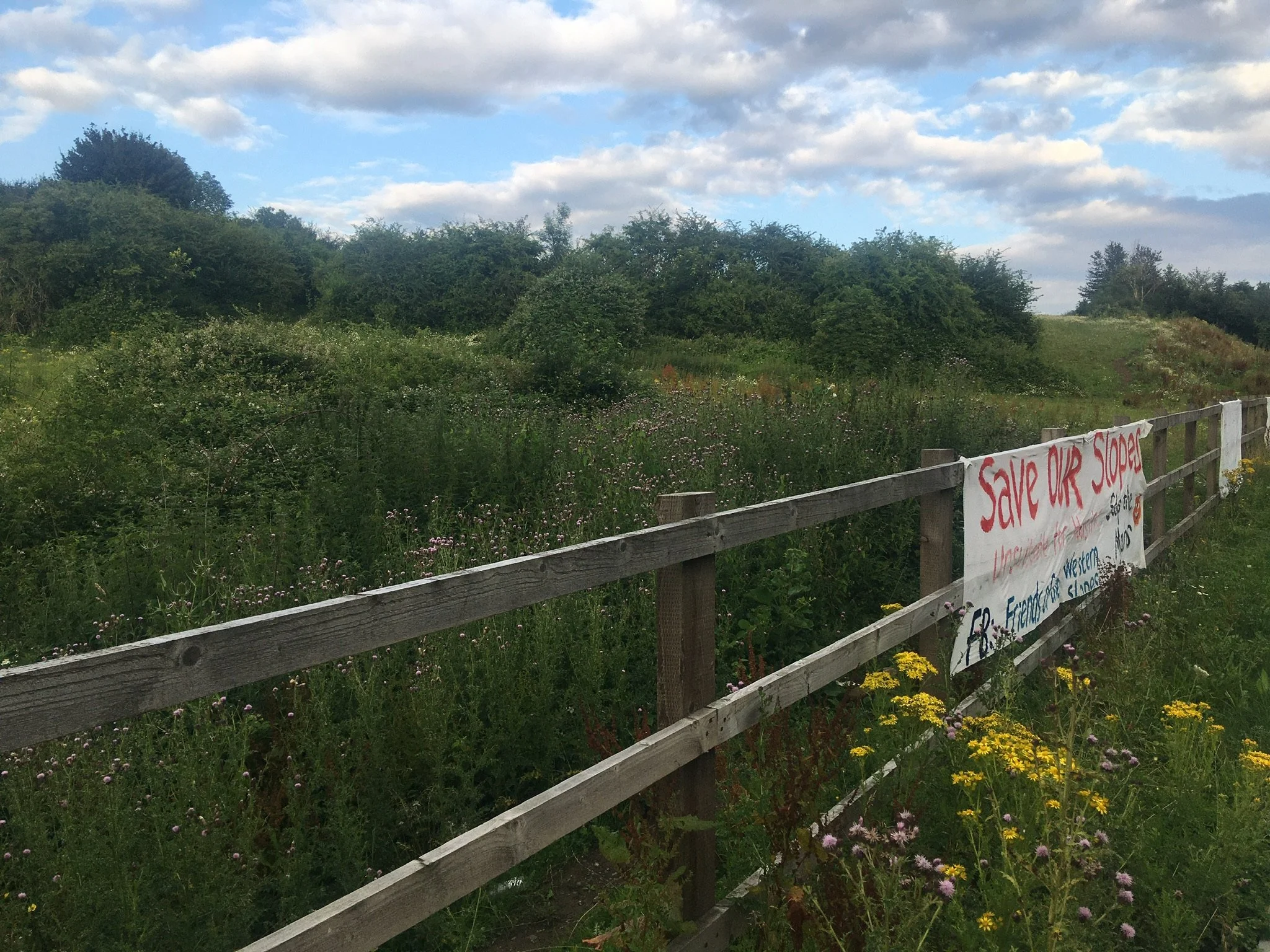 A fence in front of a field. A banner hangs off the fence reading 'Save our slopes'