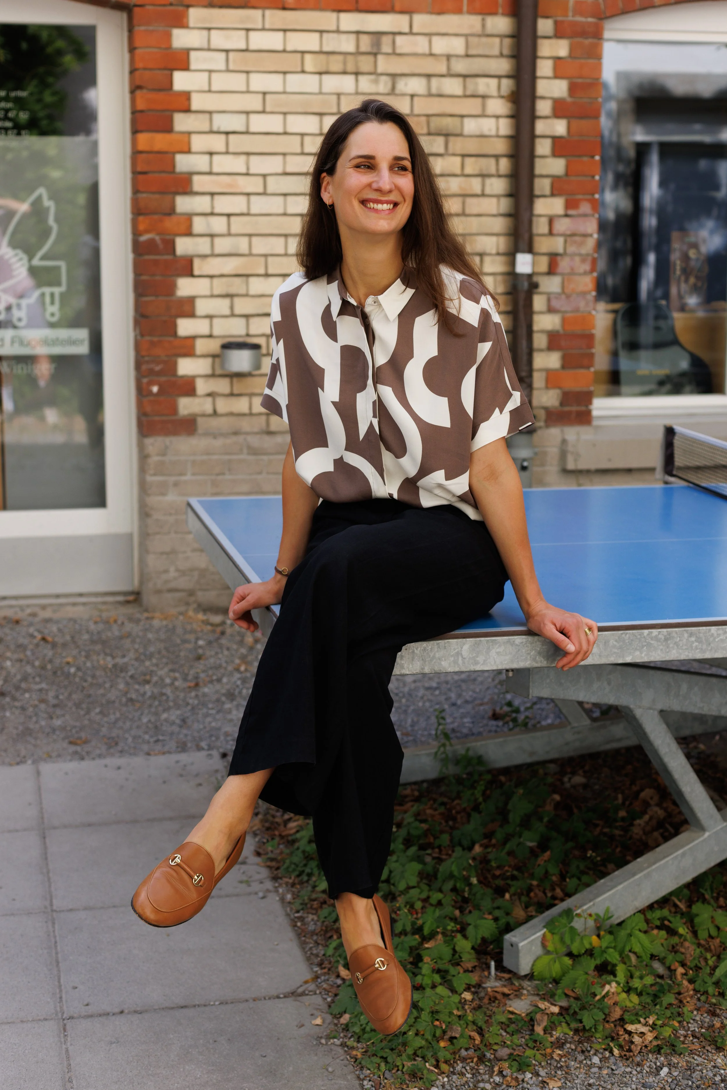 Eine lachende Frau sitzt an einem Tisch im Freien vor einem Backsteinhaus, trägt eine Bluse mit braun-weißen Mustern, schwarze Hose und braune Loafer. Sie sitzt entspannt mit einem Bein auf dem Tisch, während ihre linke Hand auf dem Tisch ruht.