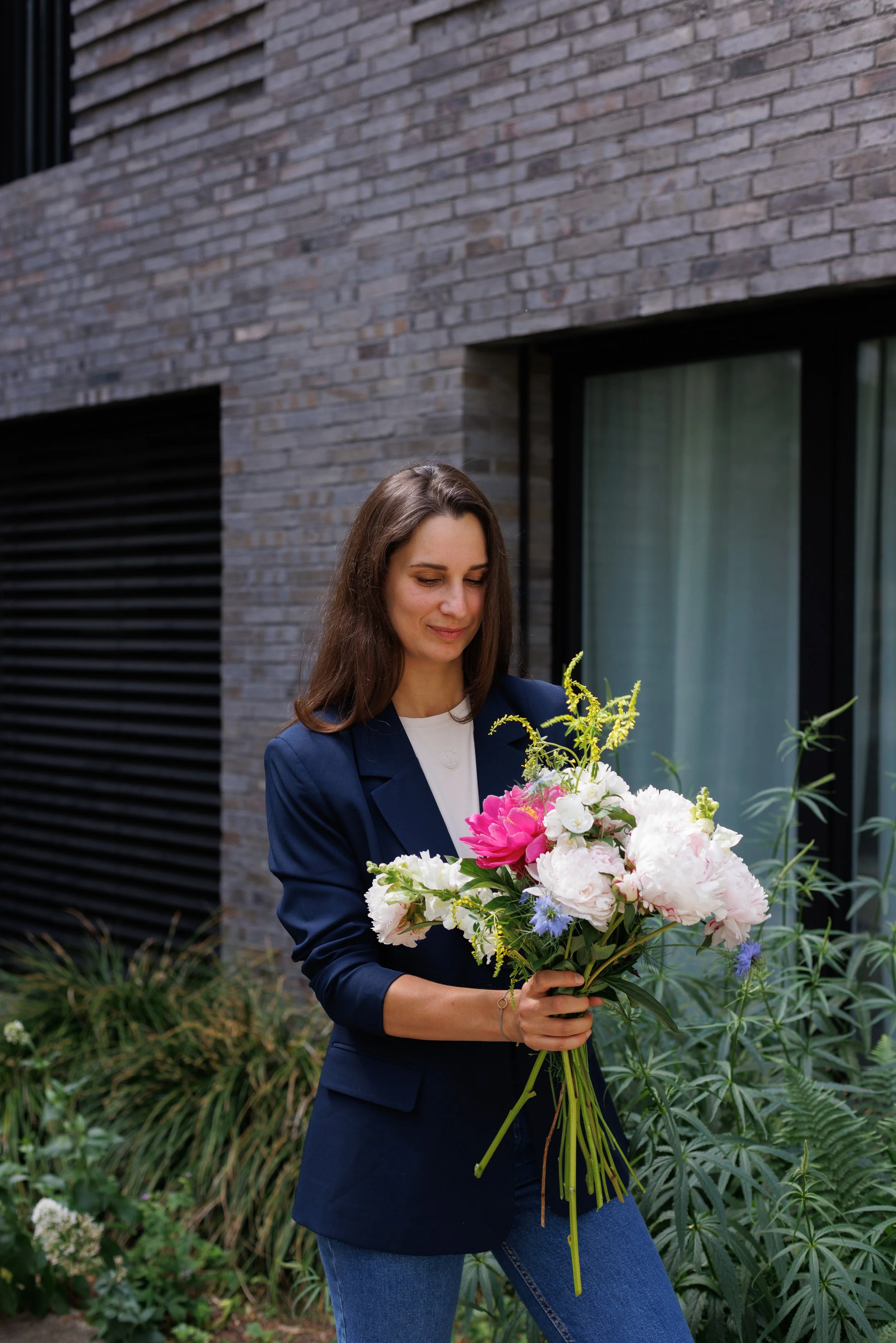 Eine junge Frau mit braunen Haaren hält einen großen Strauß bunter Blumen in den Händen. Sie trägt eine dunkle Blazerjacke und blaue Jeans. Im Hintergrund sieht man eine moderne Hausfassade mit grauen Ziegelsteinen und Fenstern.